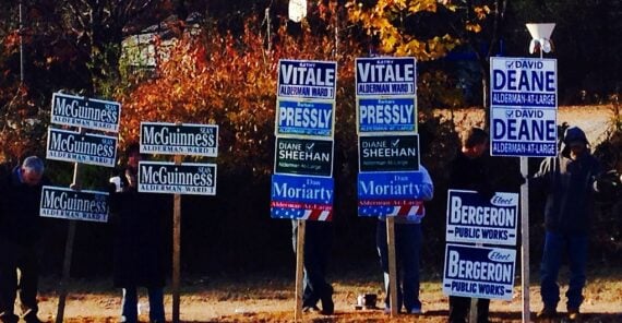 A group of people stand by the roadside holding multiple campaign signs for various local candidates, including McGuinness, Vitale, Pressly, Sheehan, Moriarty, Deane, and Bergeron, with autumn trees in the background.