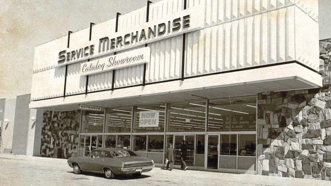 A vintage photo shows the exterior of a Service Merchandise store with “Catalog Showroom” on the sign, an old car parked in front, and people entering through glass doors.
