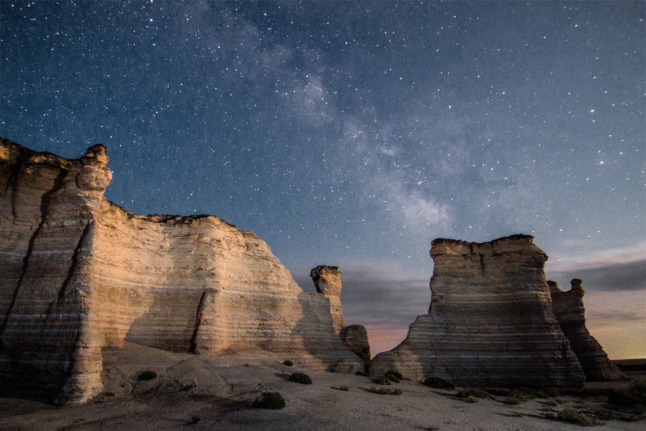 Tall rock formations under a clear night sky filled with stars and the Milky Way, with soft shadows and sparse vegetation on the ground.