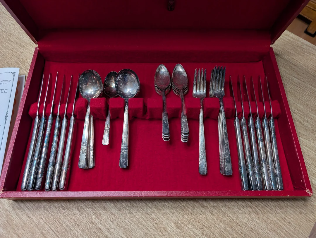 A red velvet-lined box holds a set of vintage silverware, including six forks, three large spoons, three small spoons, and six smaller forks, neatly arranged on a wooden surface.