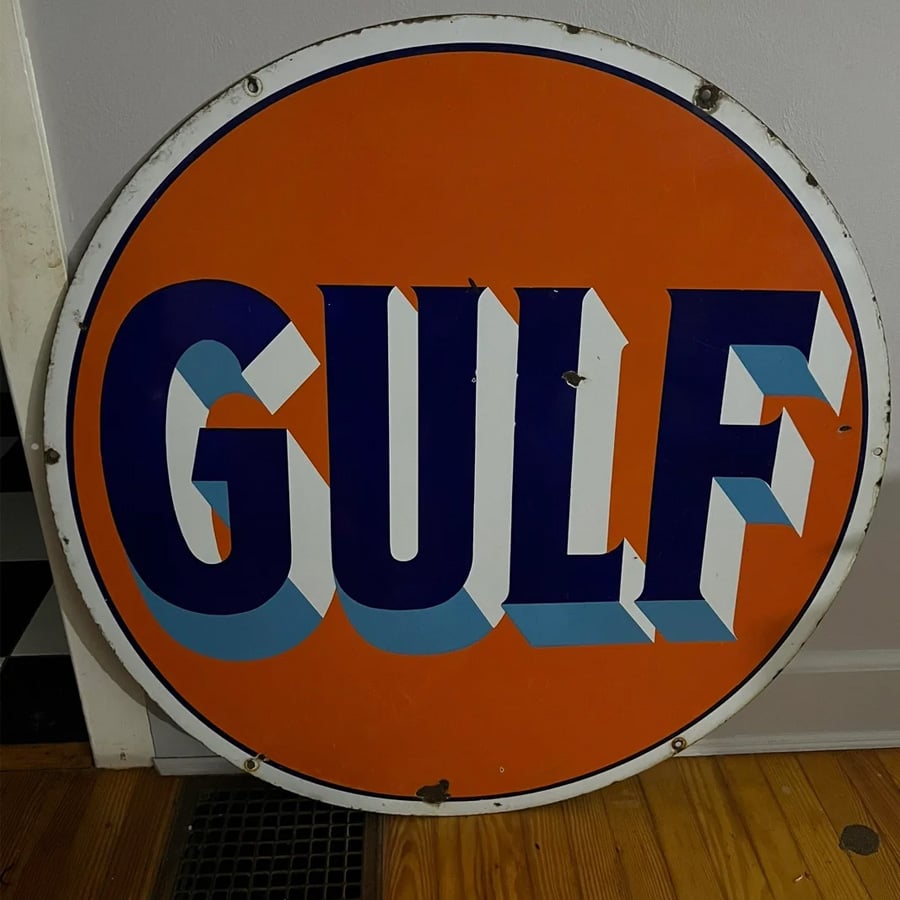 Round vintage metal sign with an orange background and bold blue and white letters spelling “GULF.” The sign shows some rust and wear, and is leaning against a wall on a wooden floor.