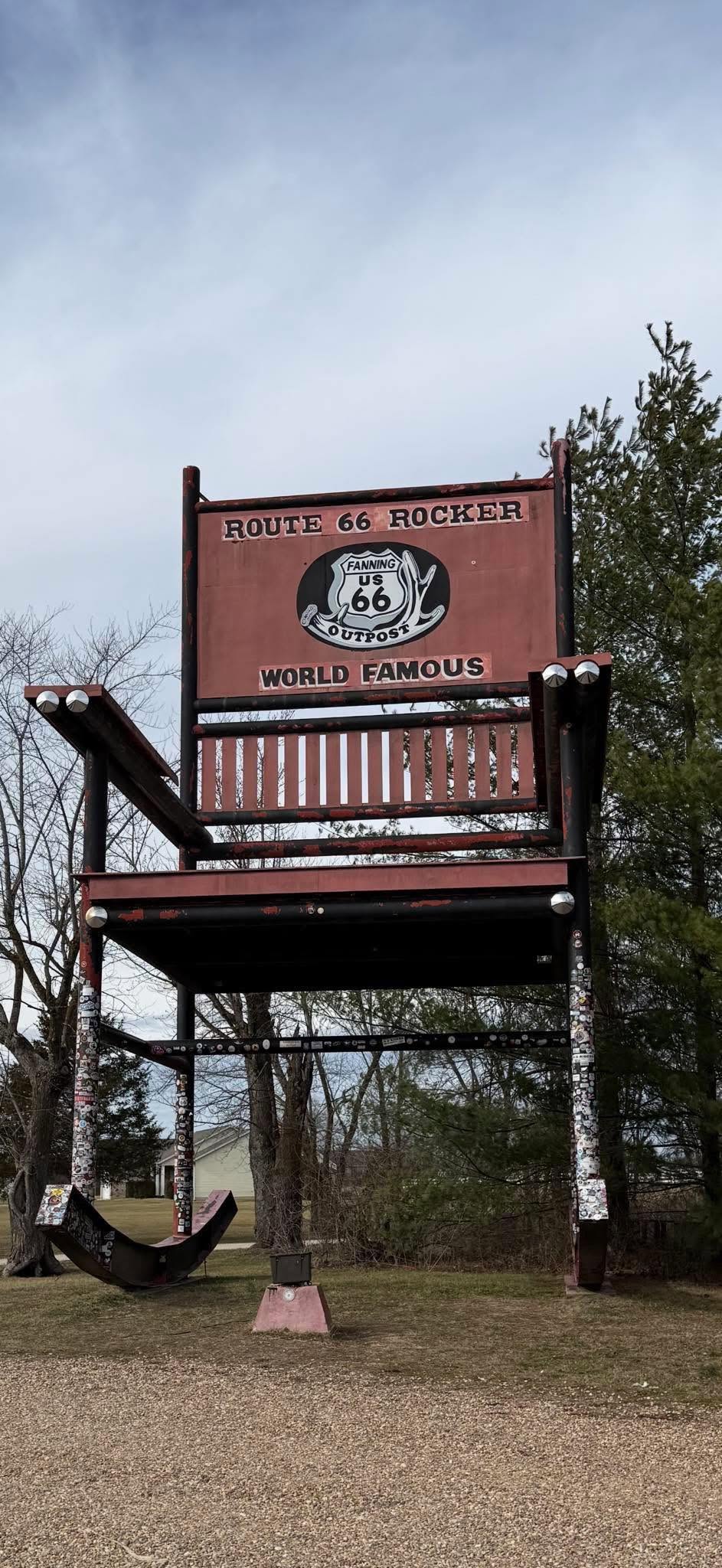 A giant rocking chair labeled “Route 66 Rocker World Famous” stands outdoors near trees and a cloudy sky, serving as a roadside attraction.