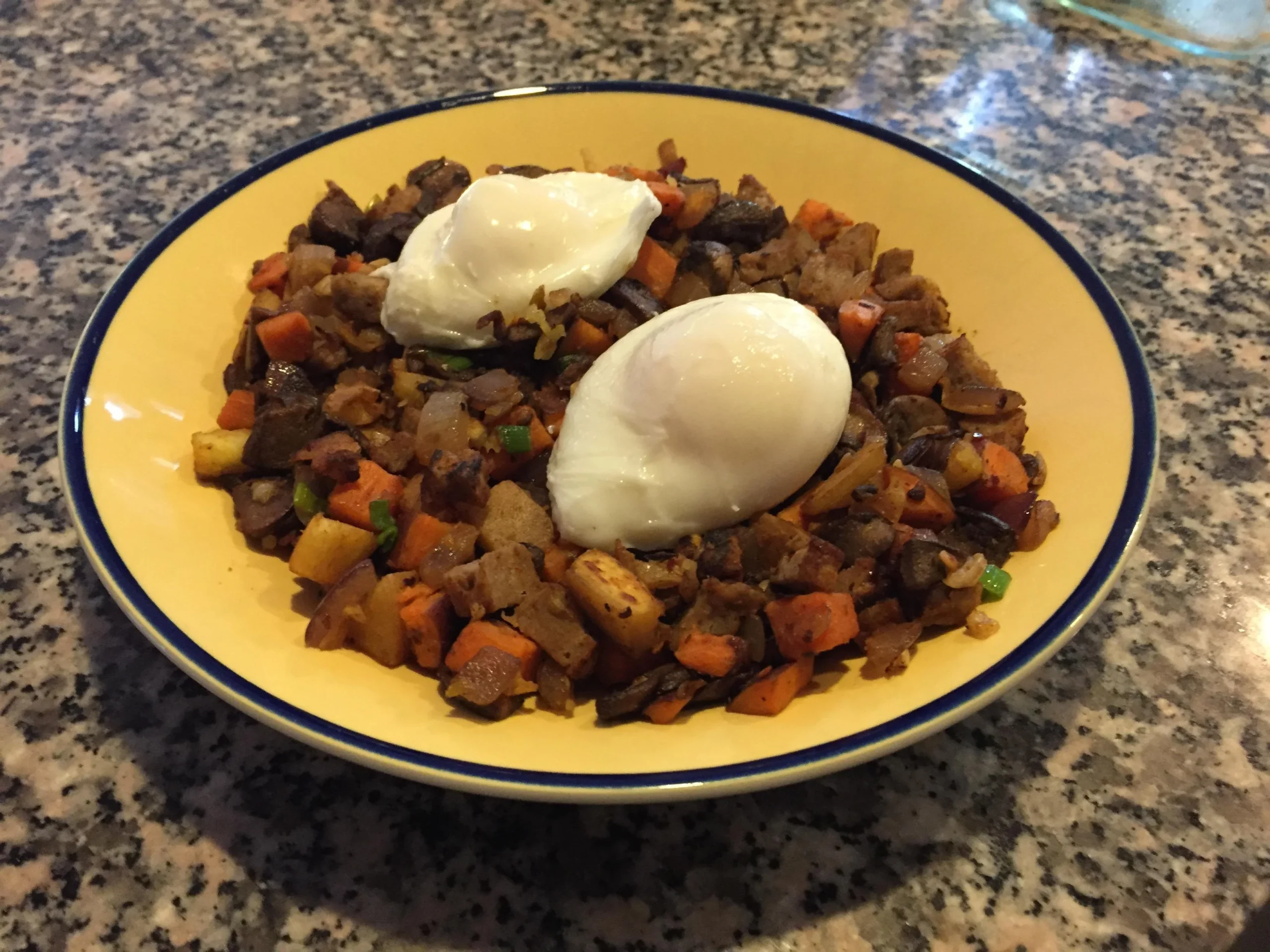 A yellow bowl filled with a hash of diced vegetables and meat, topped with two poached eggs, sits on a granite countertop.