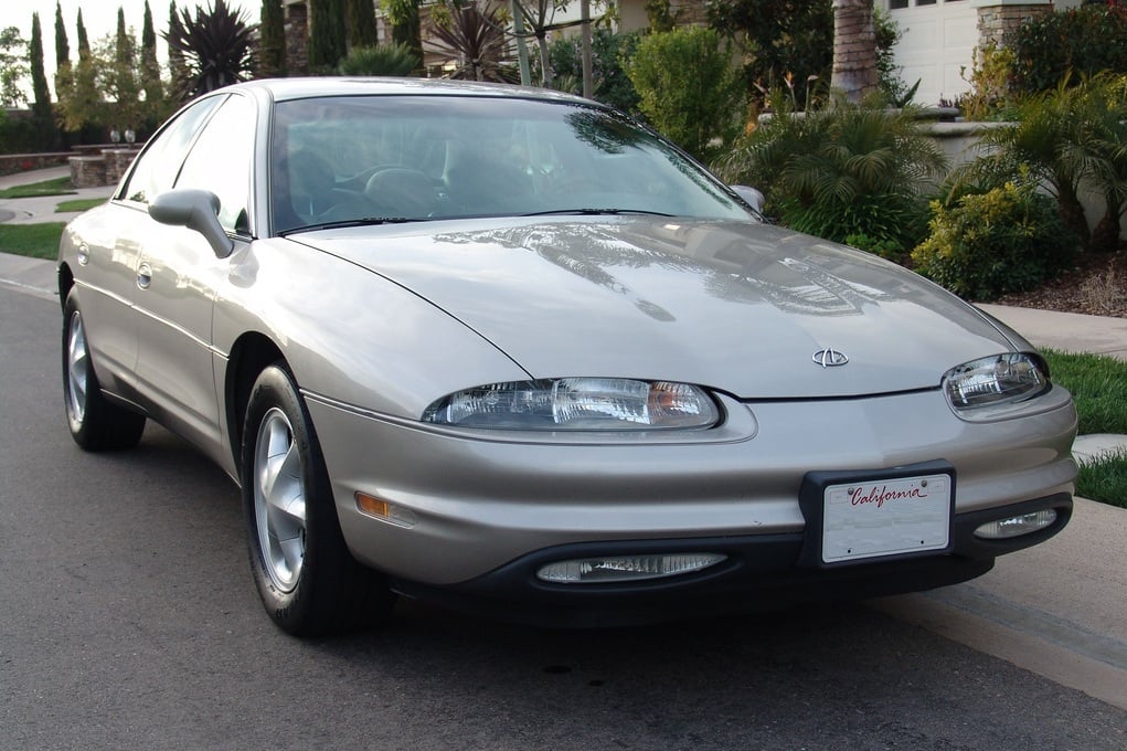 A silver two-door car is parked on a suburban street, with greenery and houses in the background. The car has a California license plate and a sleek, rounded design.
