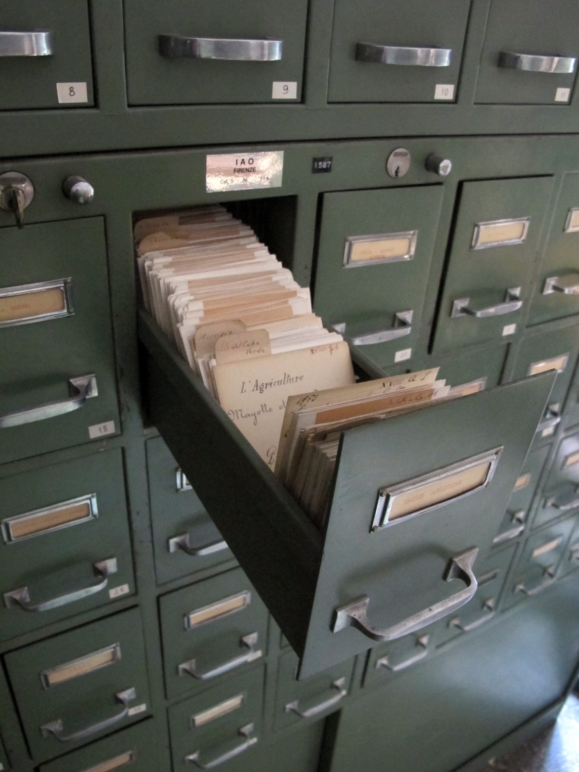 A green metal filing cabinet with one drawer open, revealing rows of handwritten index cards organized inside. Other drawers remain closed.
