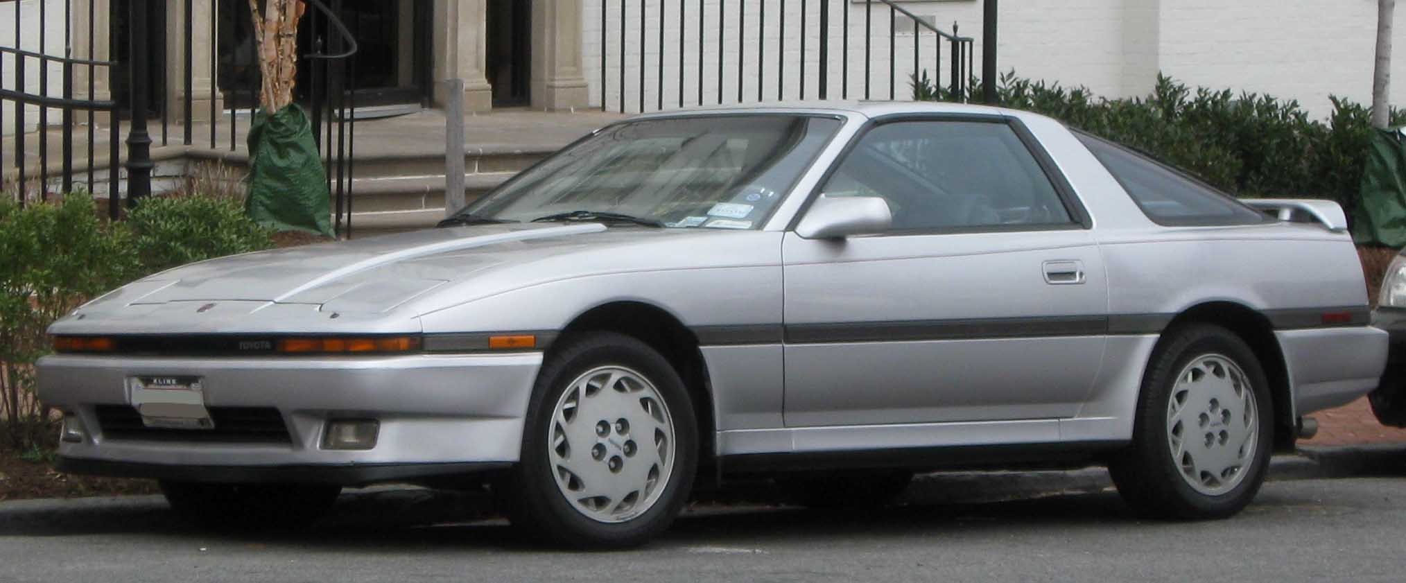 A silver sports car with pop-up headlights and circular alloy wheels is parked on a street in front of a building with steps and railings.