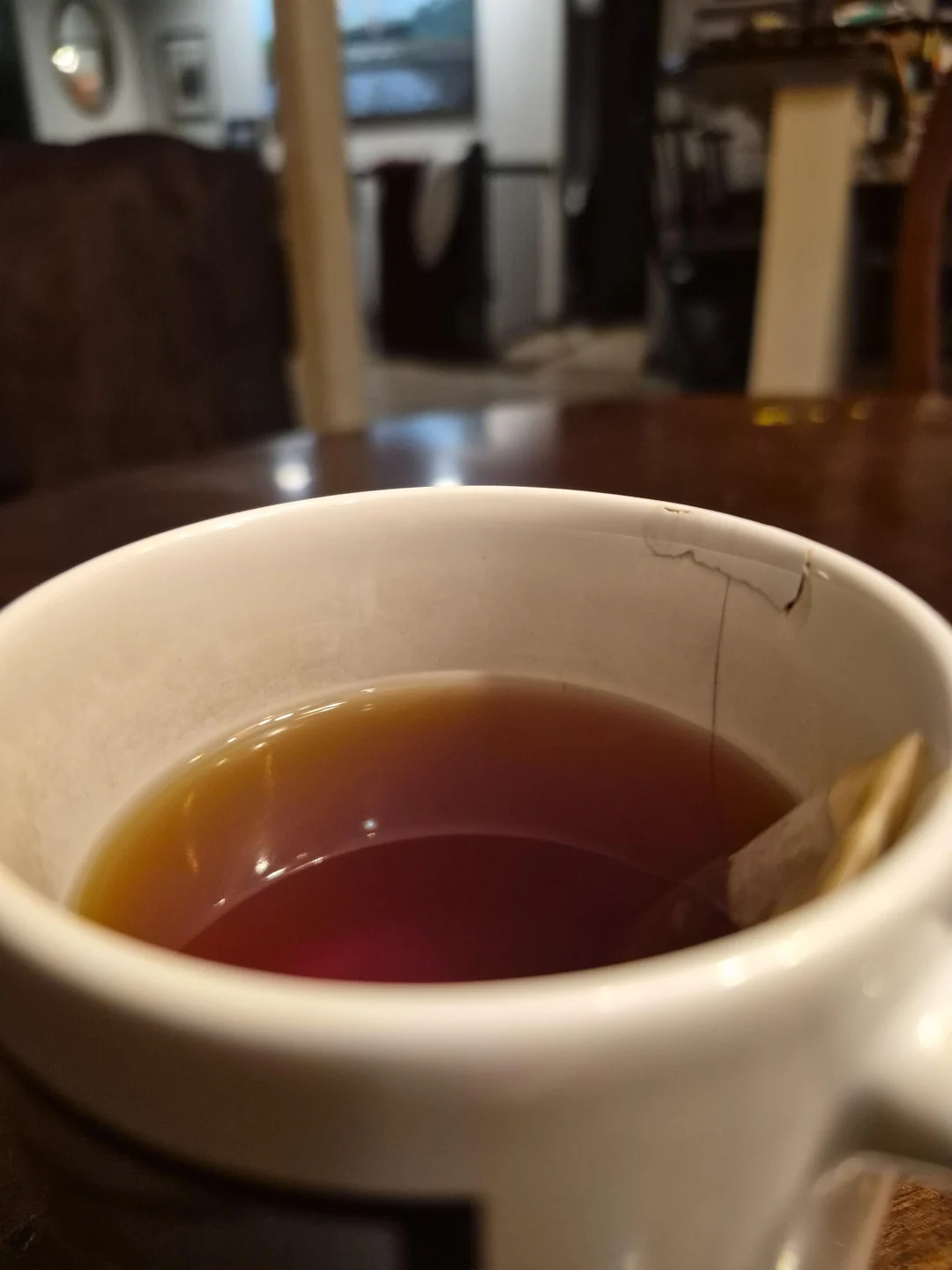 A close-up of a white mug filled with tea, with a tea bag string on the edge. There is a visible crack on the rim of the mug, and a dimly lit room is blurred in the background.