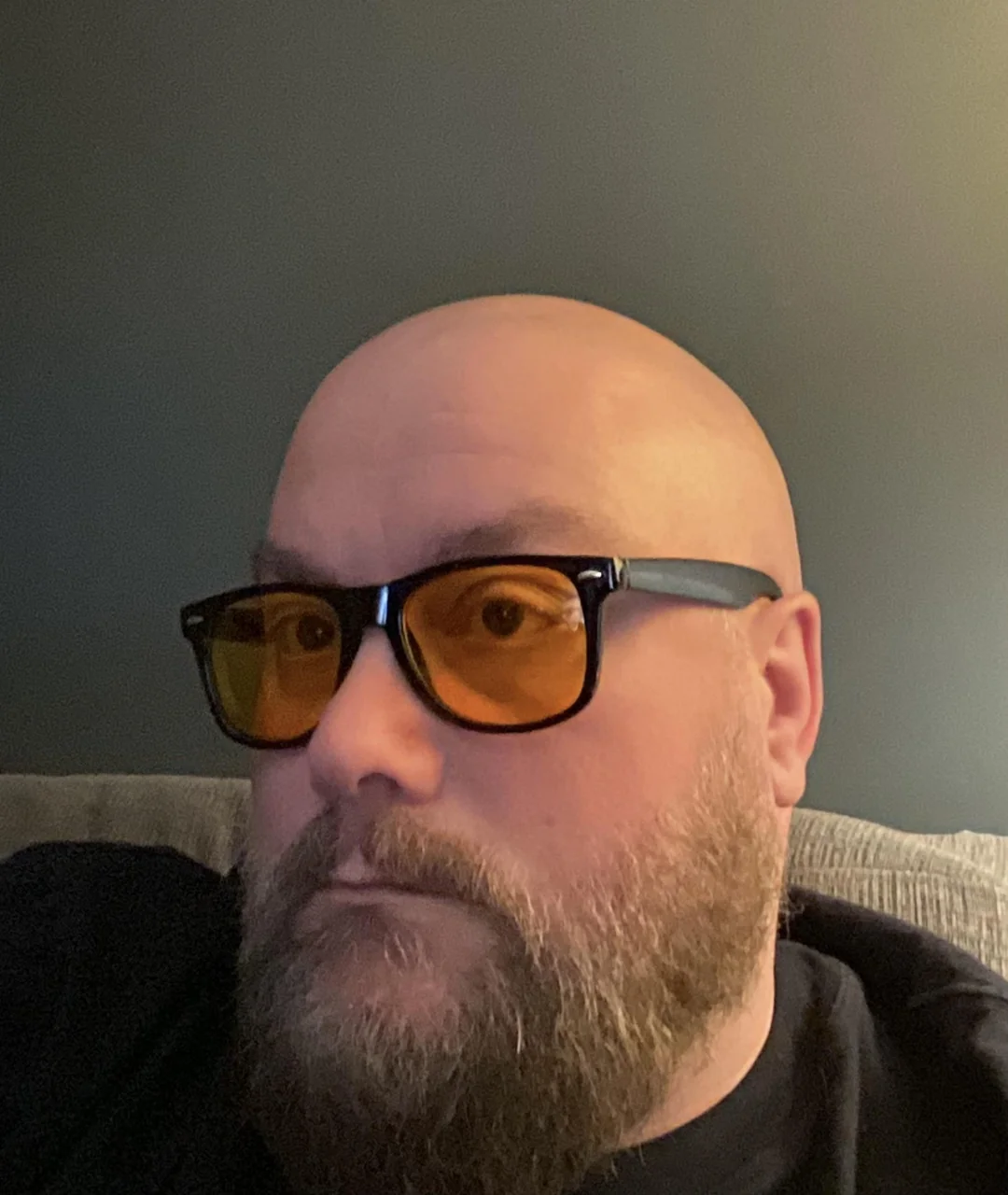 A bearded person with a bald head wears black-framed glasses with yellow lenses, sitting indoors against a gray wall, looking slightly away from the camera.