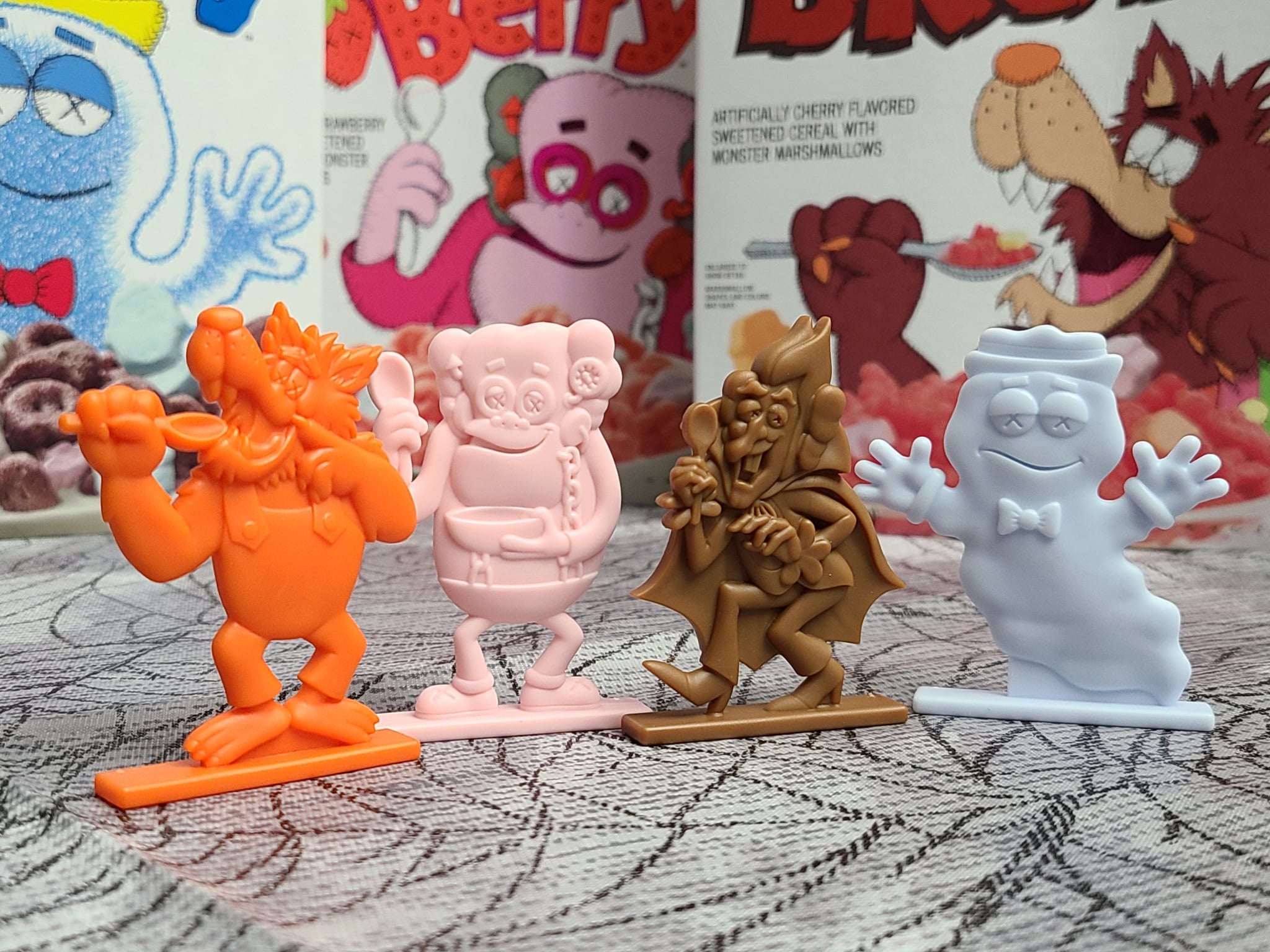 Four colorful plastic toy figures of cereal mascots stand in a row on a table, with vintage cereal boxes featuring illustrated characters in the background.
