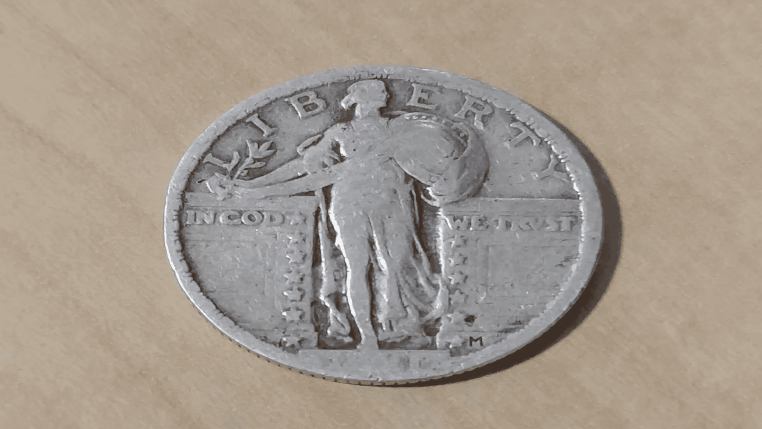 A worn silver coin depicting a standing figure holding a shield and olive branch, with “LIBERTY” above and “IN GOD WE TRUST” inscribed. The background is a light wood surface.