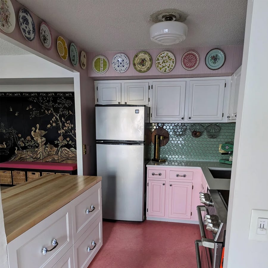 A small kitchen with pink lower cabinets, white upper cabinets, a stainless steel fridge, hexagonal green tile backsplash, and decorative plates displayed along the top wall.