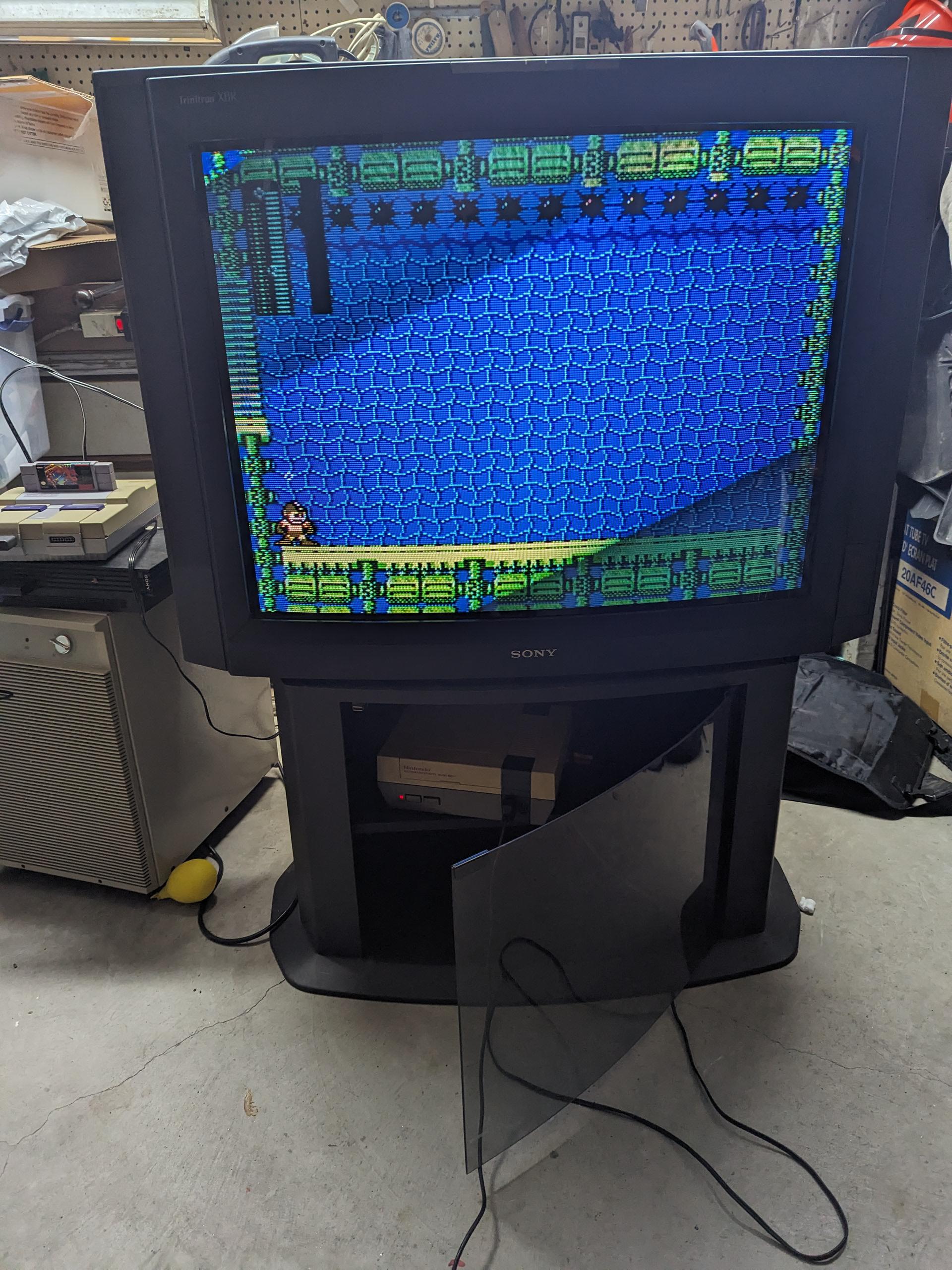 A retro video game is displayed on a CRT Sony TV. The TV stand door is open, showing a Nintendo console inside. Various electronics and tools are scattered around in a cluttered garage or workshop.