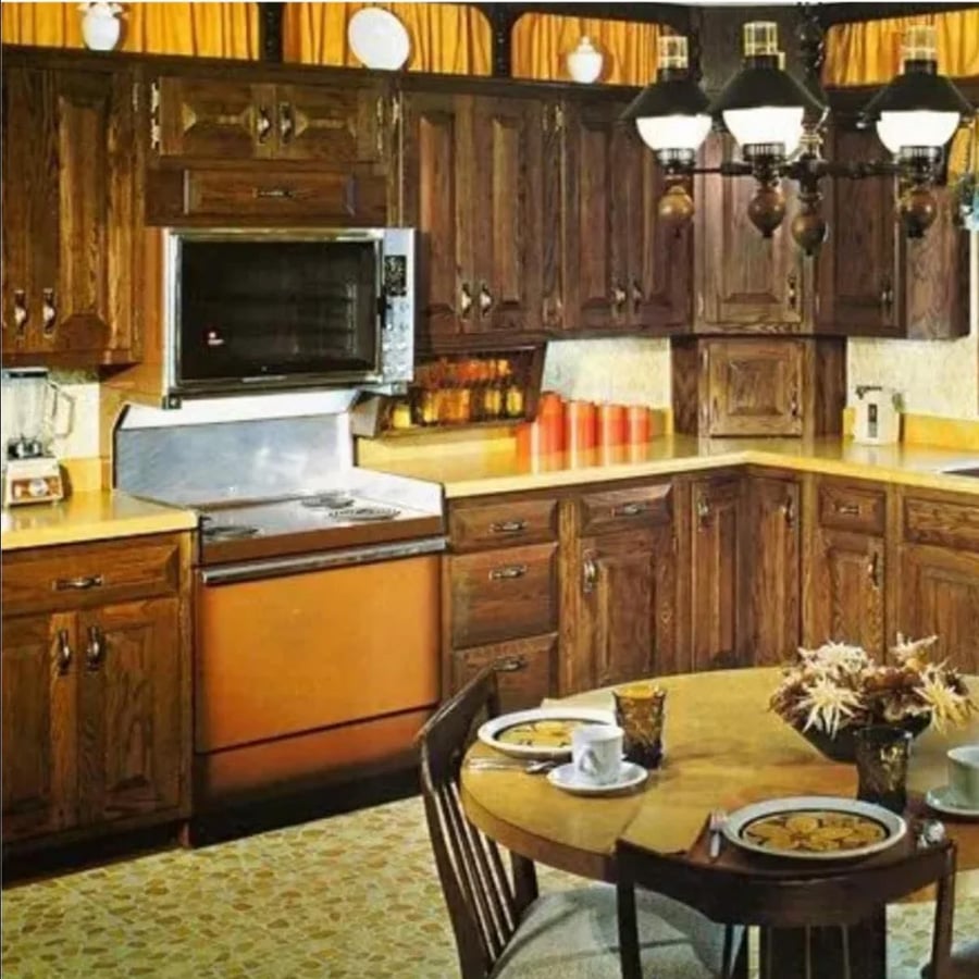 1970s-style kitchen with dark wood cabinets, gold appliances, yellow countertops, patterned floor, hanging lights, and a small round table set for two with plates, coffee cups, and a floral centerpiece.