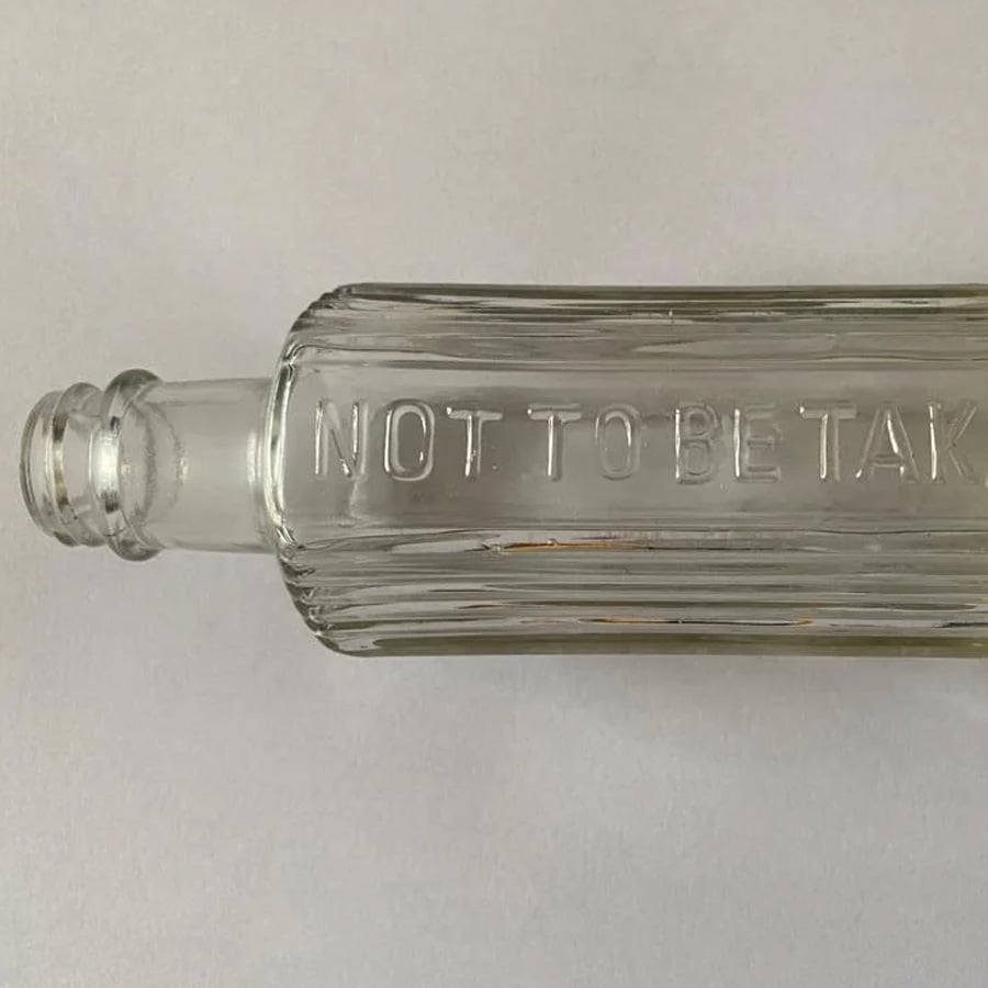 A clear, vintage glass bottle lying on its side with the raised text “NOT TO BE TAKEN” embossed on its surface. The bottle is empty and photographed on a plain white background.
