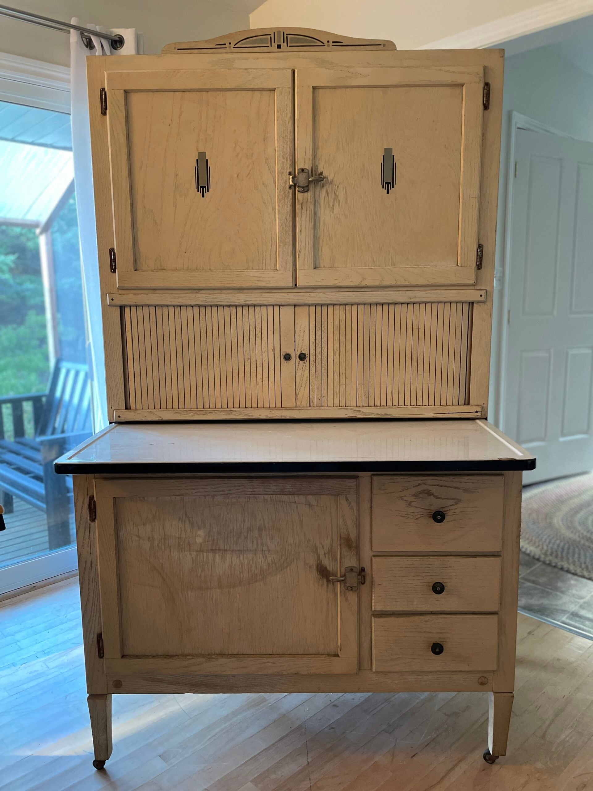 A vintage wooden Hoosier cabinet with upper and lower storage, a pull-out workspace, metal handles, and a tambour door, situated in a bright room with a window and visible greenery outside.