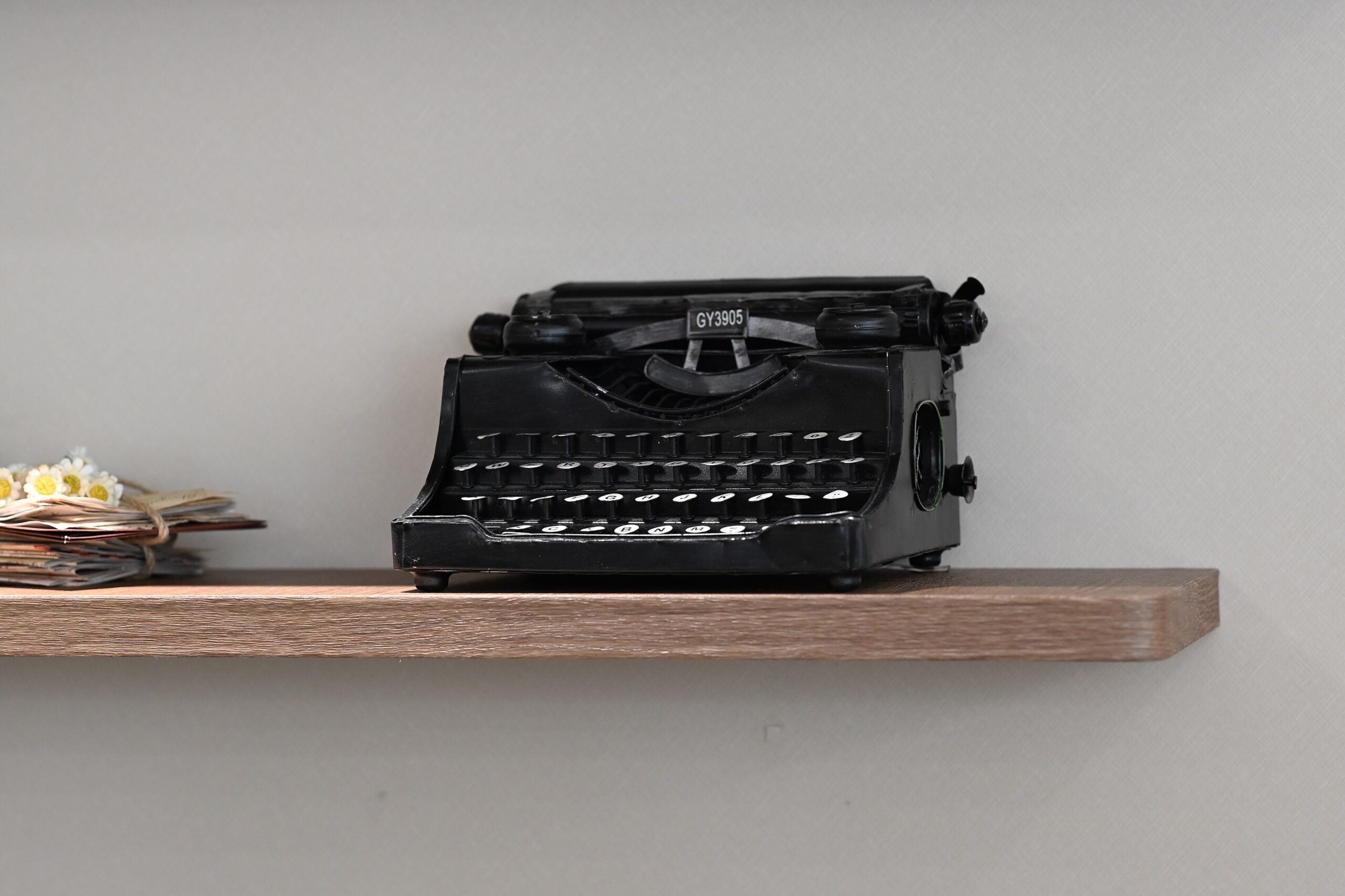 A vintage black typewriter sits on a light wooden shelf beside a small stack of papers and flowers, against a plain light gray wall.