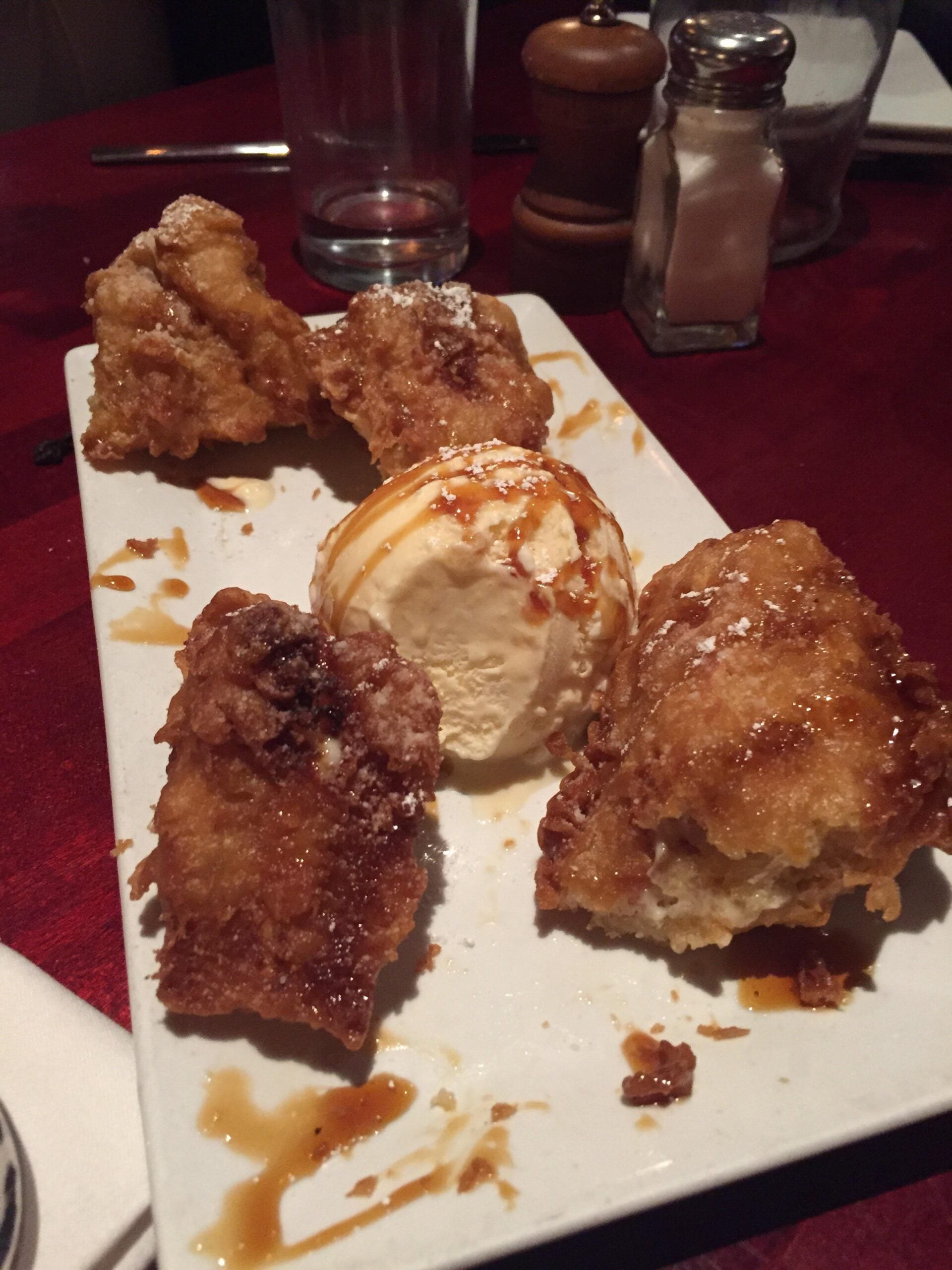 A rectangular plate with fried dessert pieces, a scoop of vanilla ice cream drizzled with caramel sauce, and a dusting of powdered sugar. There are salt and pepper shakers and an empty glass in the background.