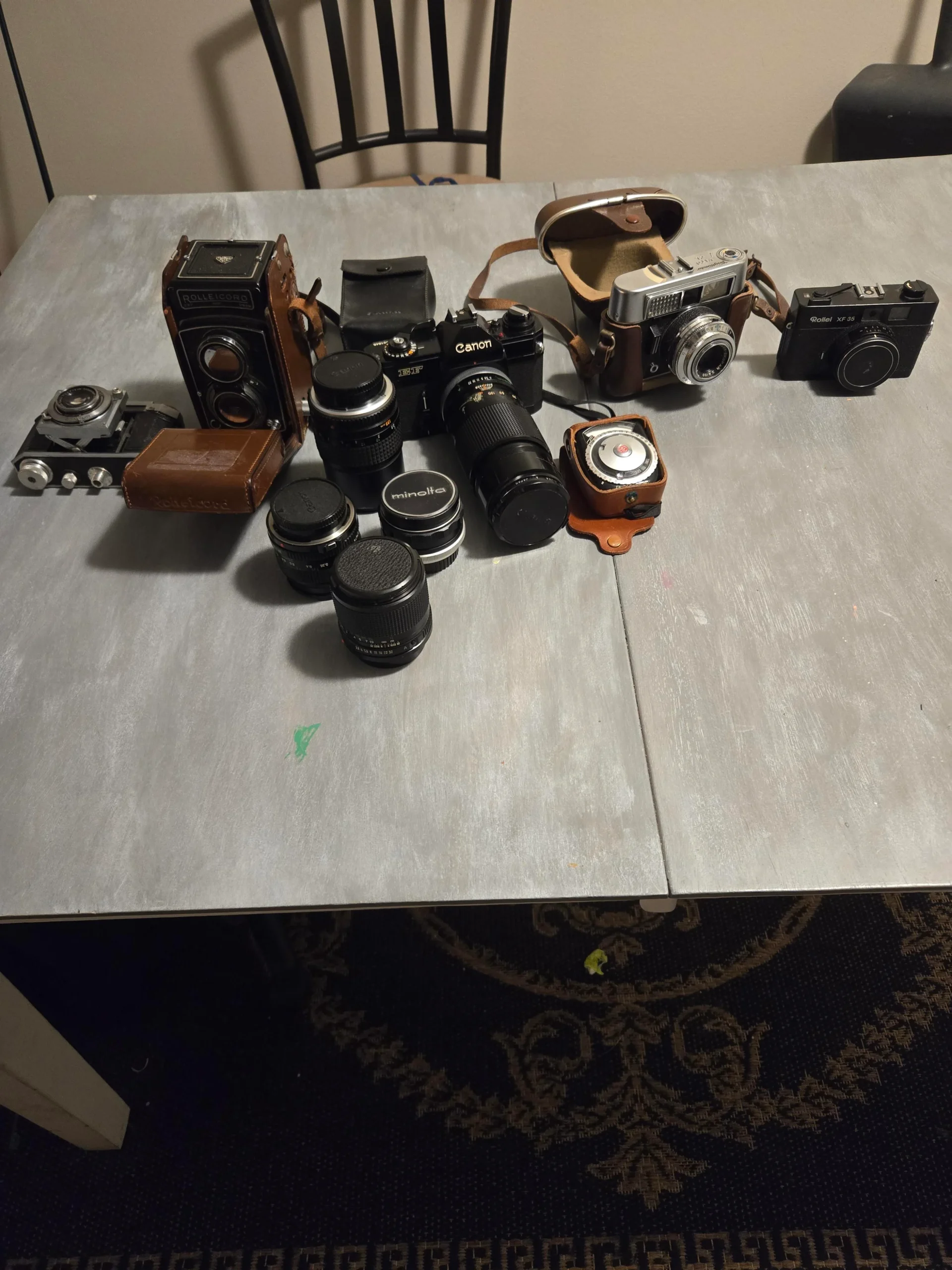 A collection of vintage cameras, camera lenses, and camera cases are arranged on a gray table with a black chair in the background.