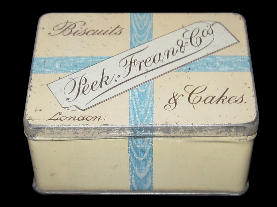 A vintage rectangular tin box labeled “Peek, Frean & Co’s Biscuits & Cakes, London” with a cream color and a blue ribbon design.