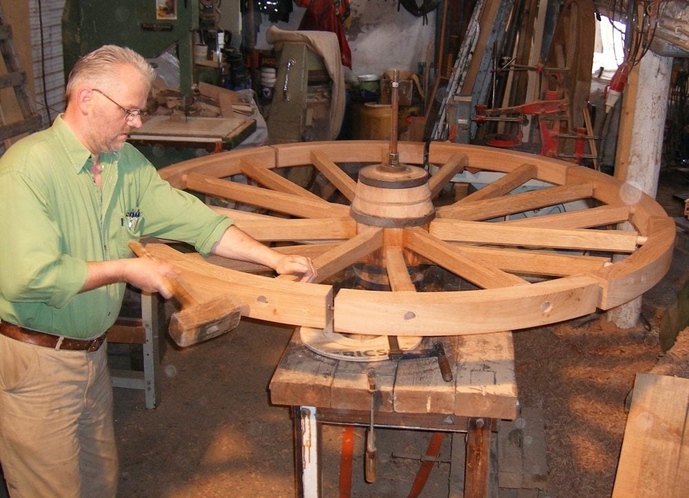 A man in a green shirt works on assembling a large wooden wagon wheel in a cluttered workshop, using tools on a workbench.