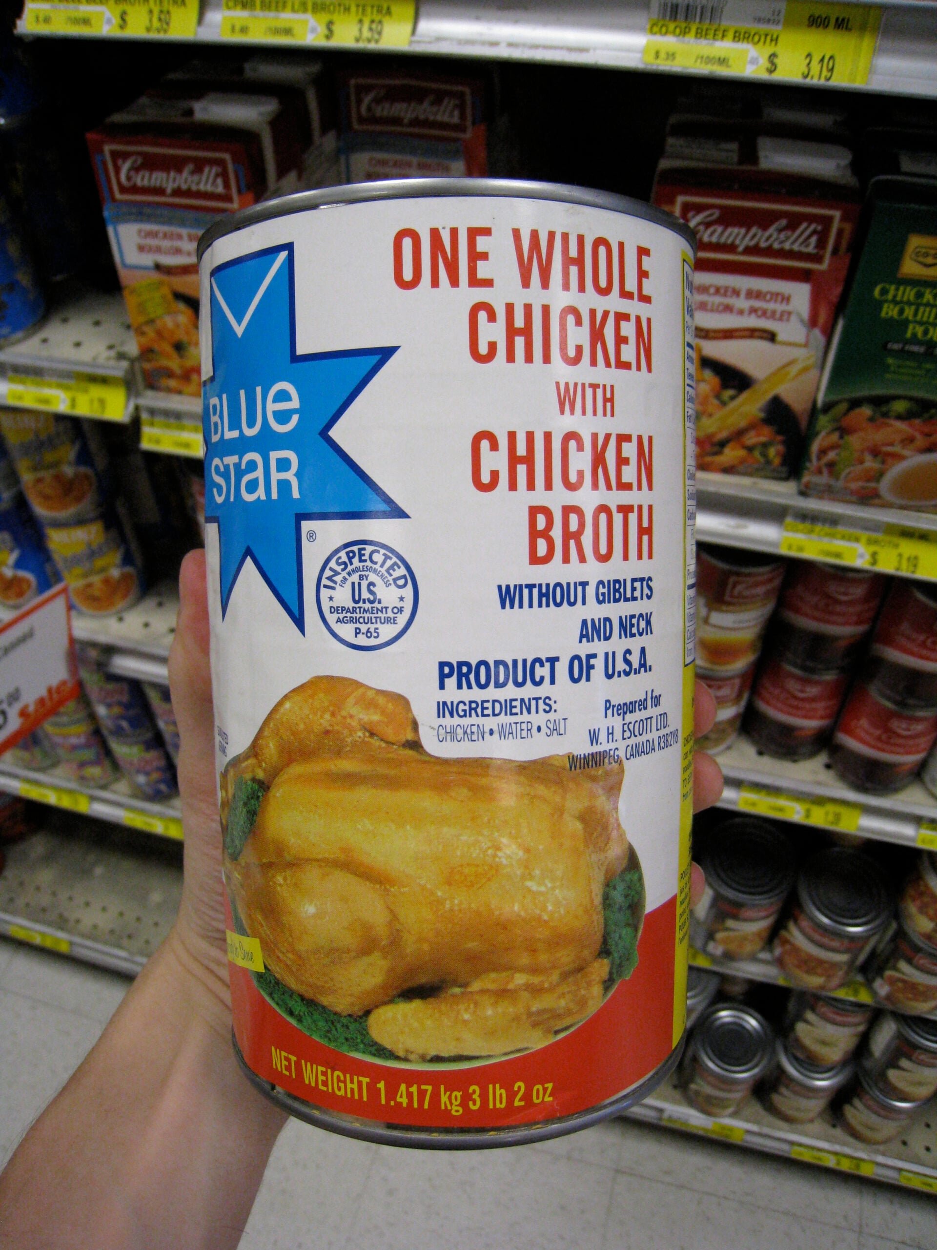 A hand holds a large can labeled “One Whole Chicken with Chicken Broth” in a grocery store aisle, with shelves of canned goods in the background. The can features an image of a cooked chicken.