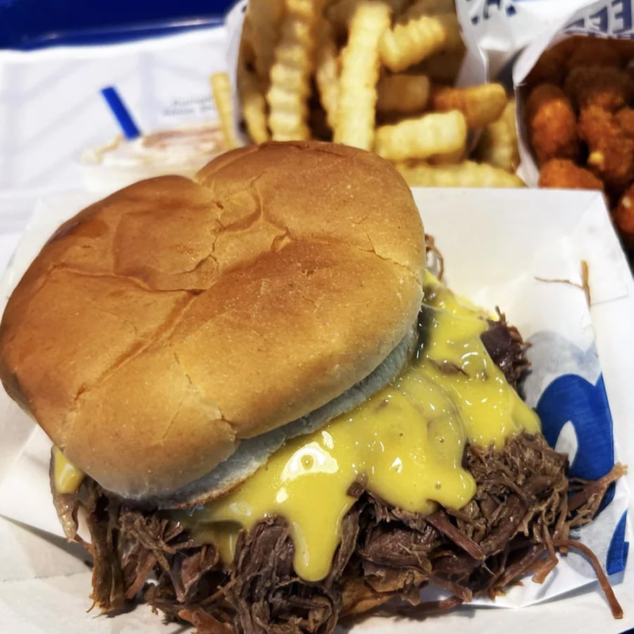 A shredded beef sandwich with yellow sauce on a bun, served with crinkle-cut fries and round fried snacks in the background.