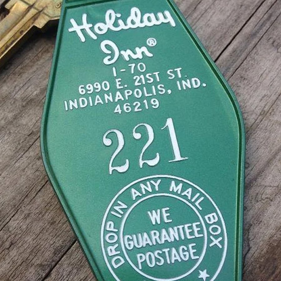 A green Holiday Inn key tag with white text showing the address, room 221, and instructions to drop in any mailbox for guaranteed postage.