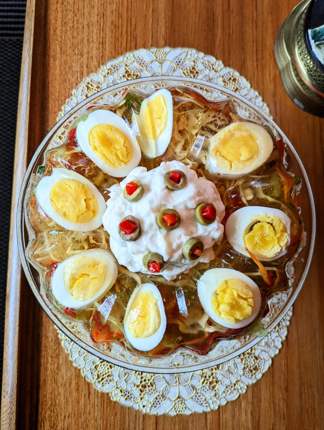 A decorative dish with sliced boiled eggs arranged around a gelatin salad, topped with a dollop of cream and chopped red chili, served on a lace doily on a wooden surface.