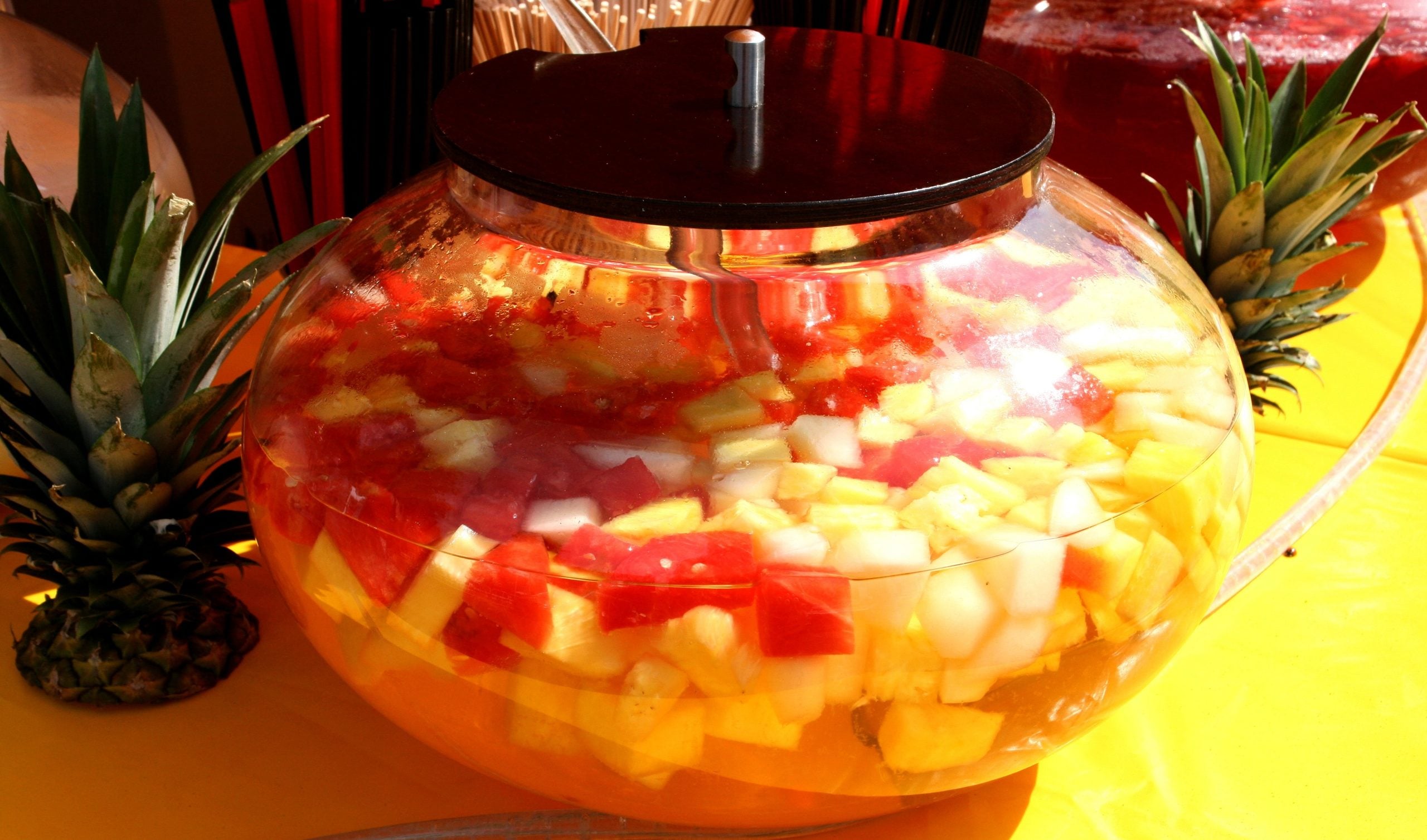 A large glass bowl contains a colorful fruit drink with chunks of pineapple, watermelon, and other fruits, placed on a yellow surface with whole pineapples nearby.