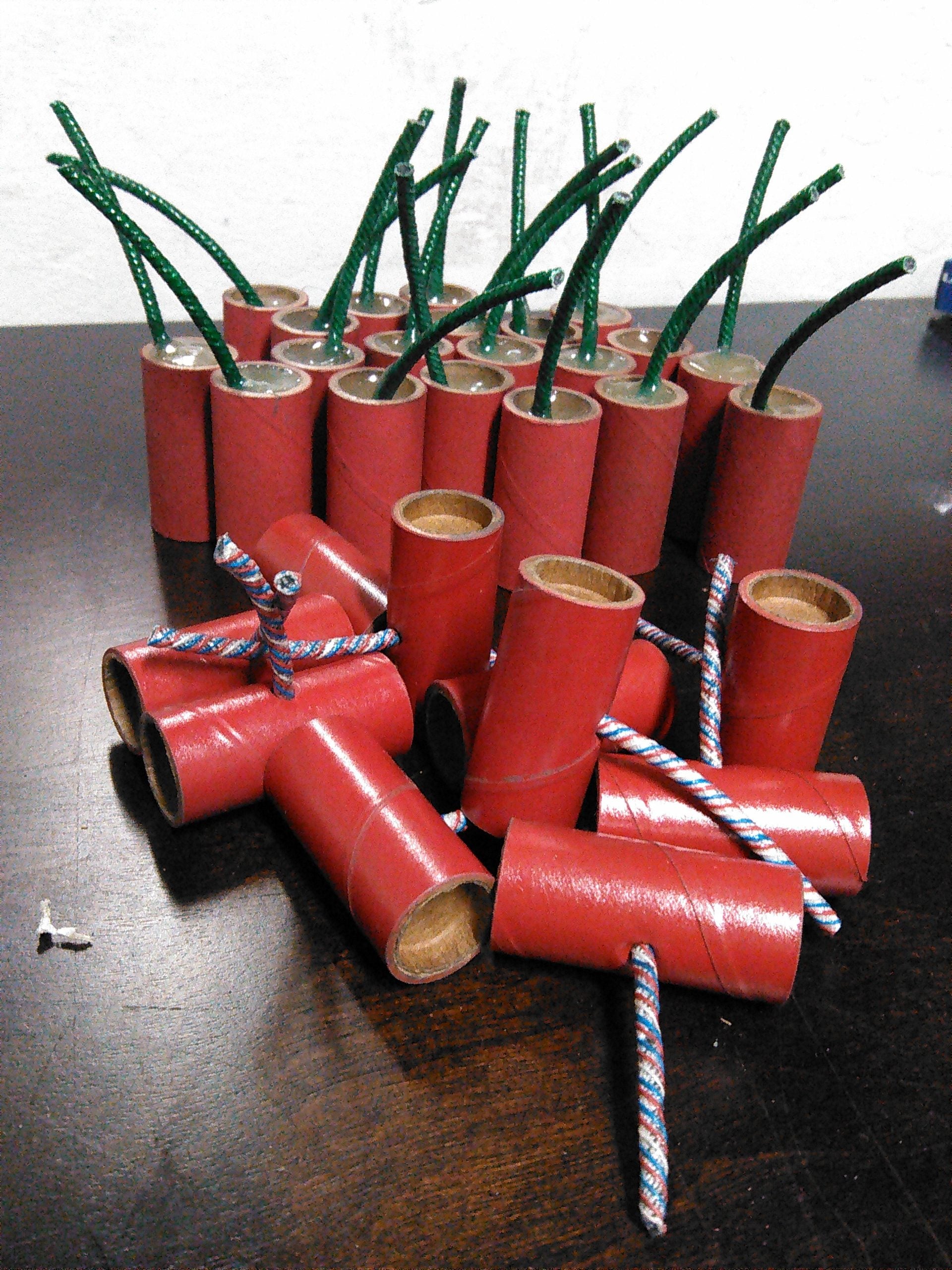 Several red firecrackers with green and blue fuses are arranged on a dark wooden surface, some stacked and some lying flat, with their fuses facing outward.