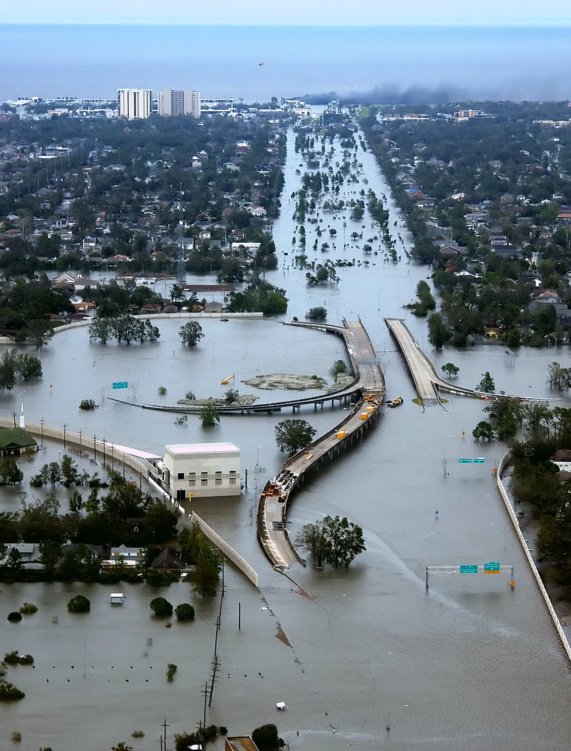 Aerial view of a flooded city with submerged roads, buildings, and vehicles, and a heavily damaged, collapsed bridge in the foreground. Water covers most of the visible area.