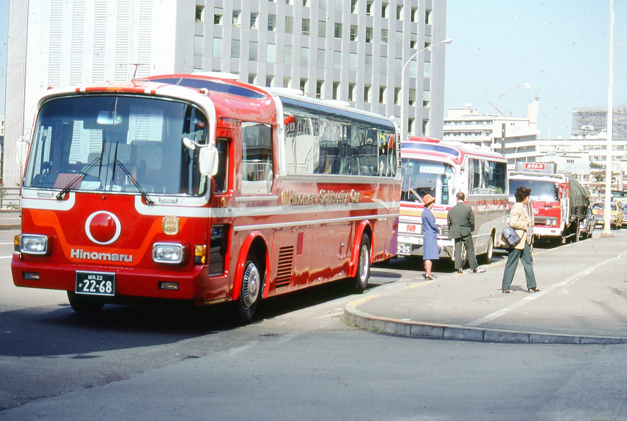 Three red and white buses are parked in a row on a city street. Several people stand near the buses. Tall buildings and urban scenery are visible in the background.