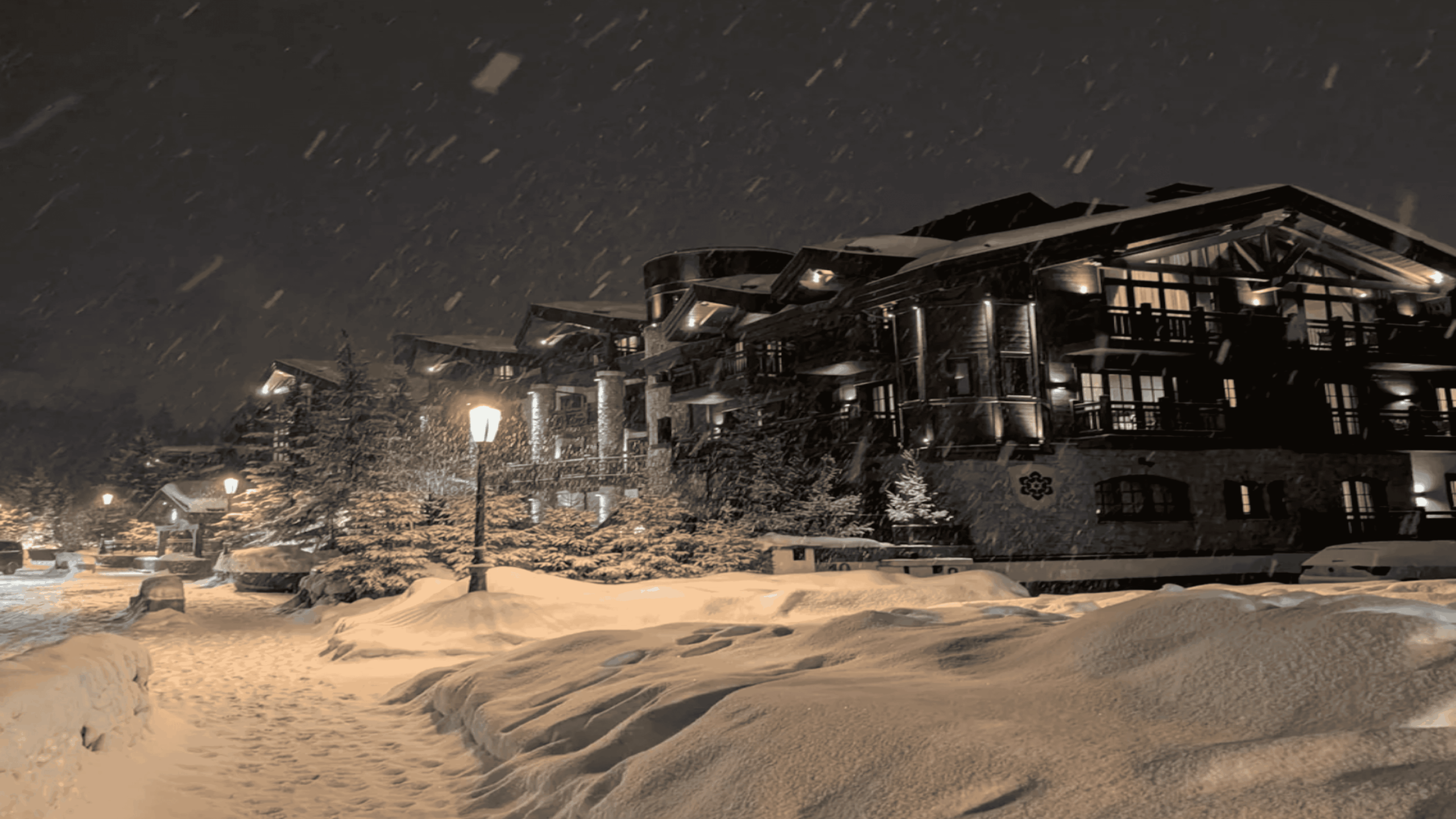 A snowy night scene with a large, warmly lit lodge surrounded by snow-covered trees and paths, as snow falls gently from the dark sky.