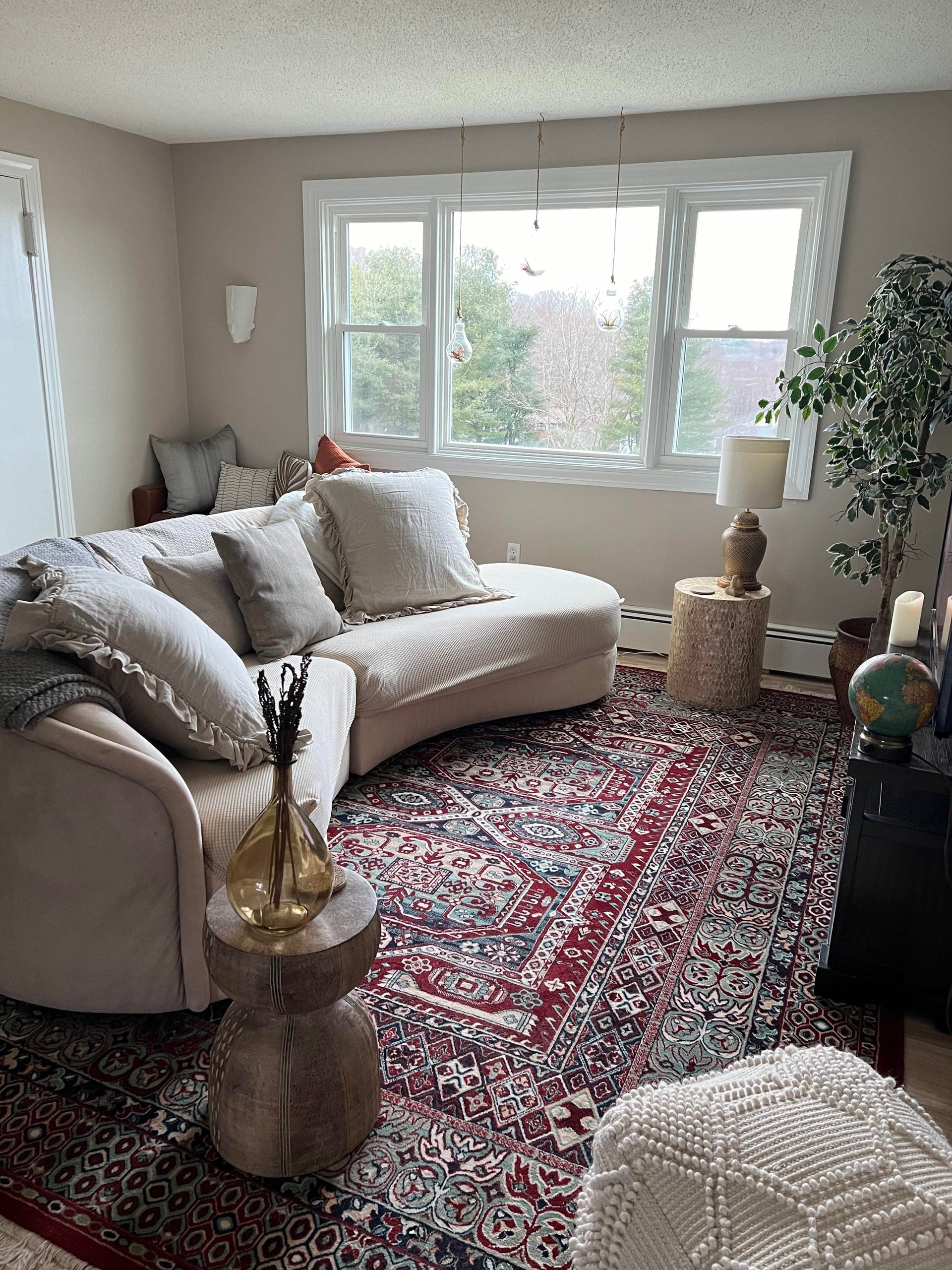 Cozy living room with a curved sofa, large pillows, colorful patterned rug, side tables, plants, a globe, candles, and large windows letting in natural light with a view of trees outside.