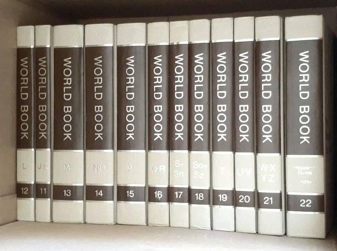 Thirteen hardcover “World Book” encyclopedia volumes, labeled with letters and numbers 12–22, are lined up on a bookshelf. The books have white spines with brown bands for the titles and numbers.