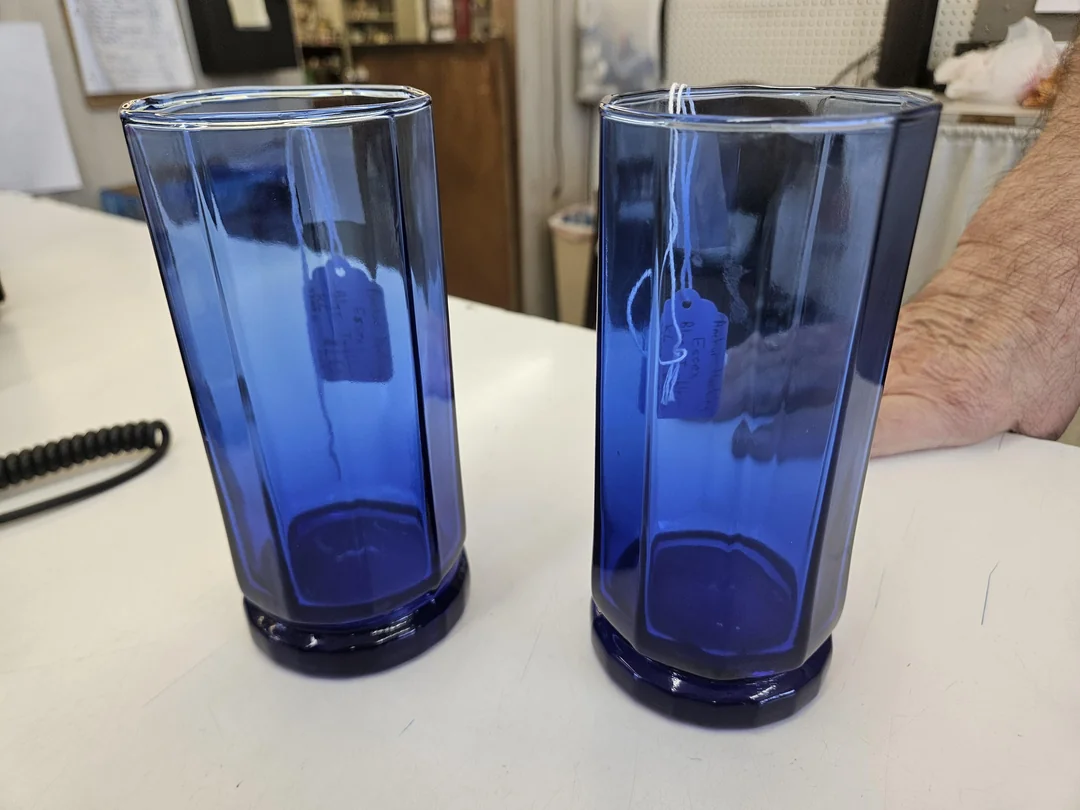 Two tall, blue glass tumblers with price tags inside are placed on a white countertop. Part of a person’s arm is visible to the right. The background shows shelves and various items.