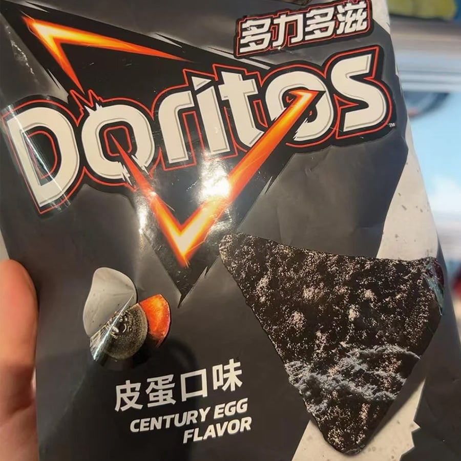 A hand holds a bag of Doritos with Chinese text. The flavor is “Century Egg,” featuring a dark chip and an image of a sliced century egg on the packaging.