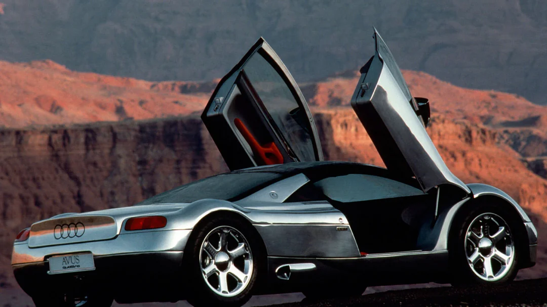 A silver Audi concept car with scissor doors open is parked in front of rocky canyon scenery under a clear sky.