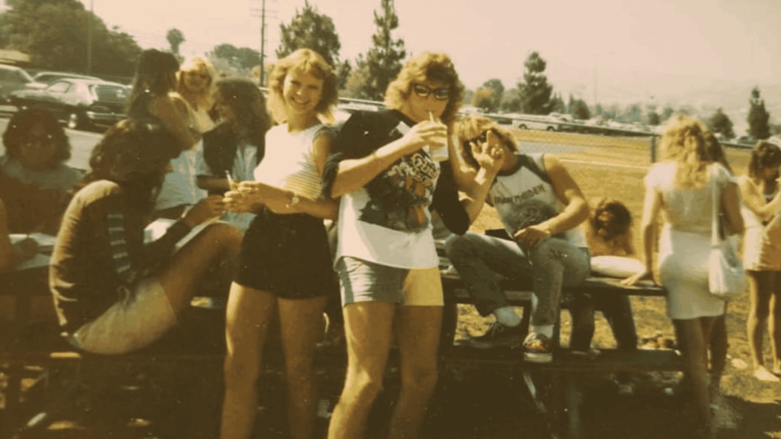 A group of teenagers hang out on and around a picnic table outdoors on a sunny day, some smiling, drinking, and talking; cars and trees are visible in the background.