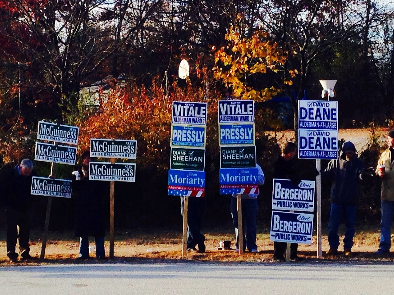 A group of people stand by a roadside holding various campaign signs for local elections, with trees and autumn foliage in the background.