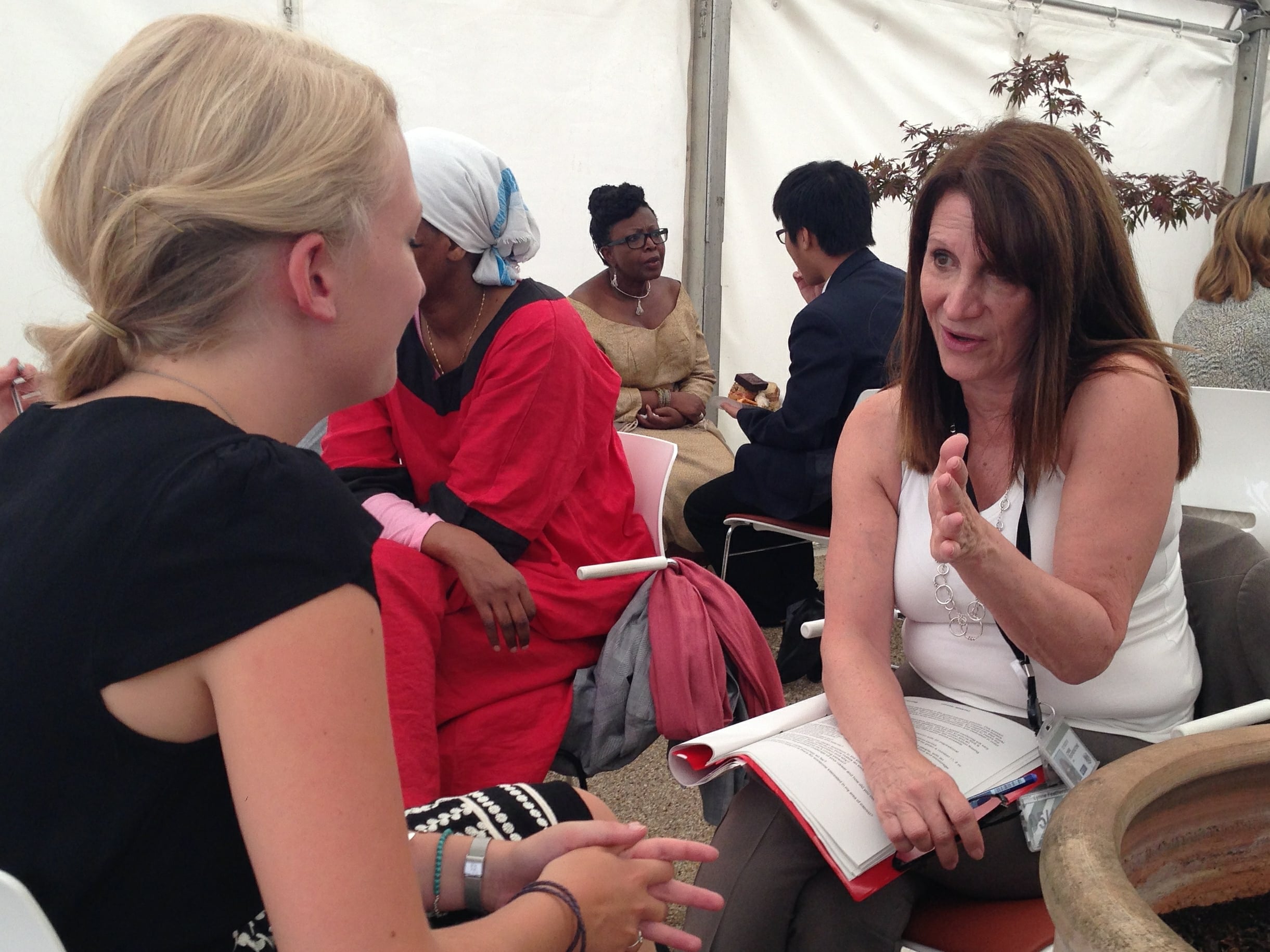 Two women sit facing each other and talking; one gestures with her hand while holding a notebook. Several other people are having conversations in the background in a bright indoor setting.