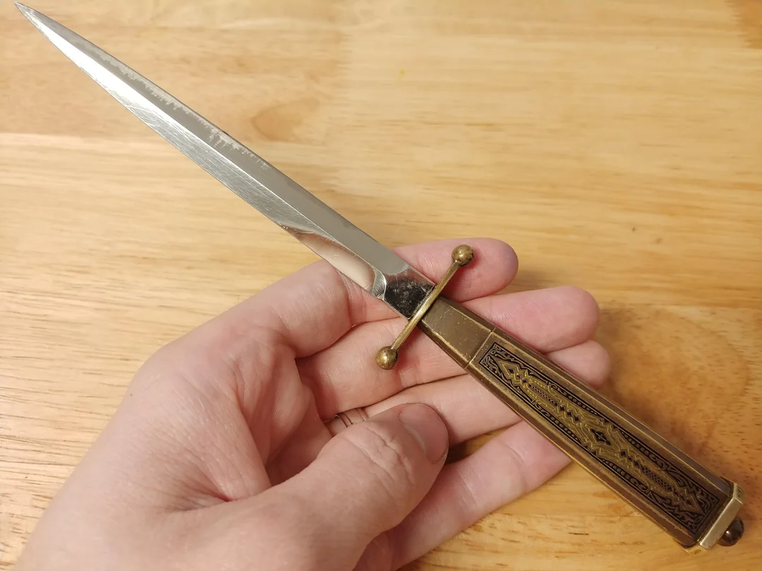 A hand holds a small, ornate dagger with a shiny silver blade and a decorated brass handle, resting on a wooden surface.