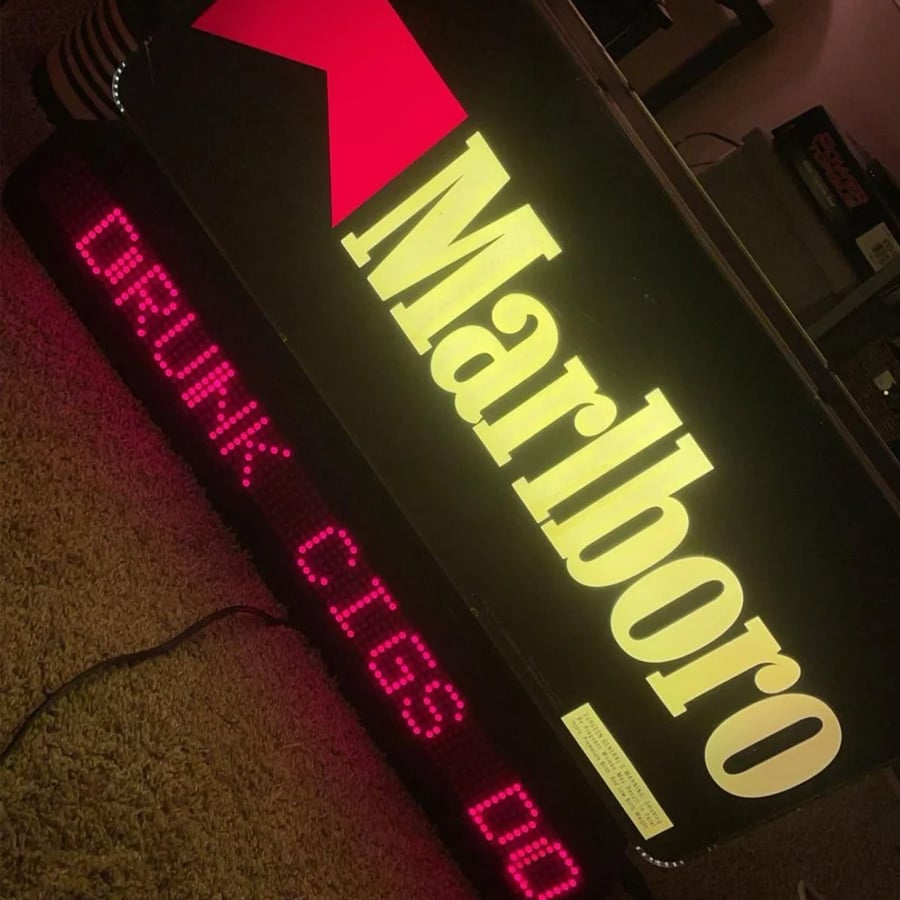 A lit Marlboro sign sits on the carpet. Below, a red LED display partially reads “DRUNK CIGS DO,” with the rest of the message cut off in the image.