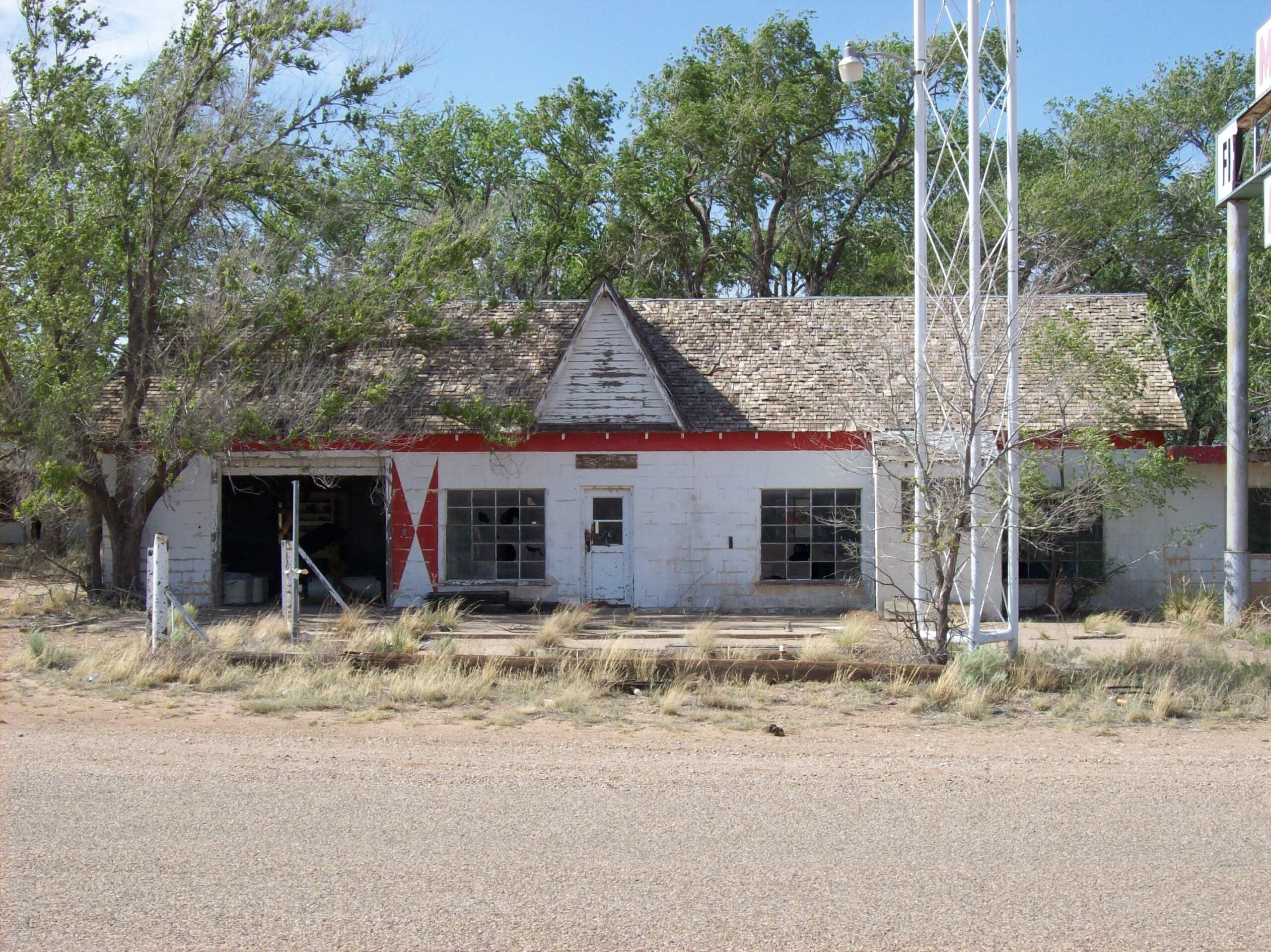 An abandoned, weathered gas station with a worn roof, broken windows, and overgrown weeds stands empty by a deserted road. Tall trees grow behind the building.