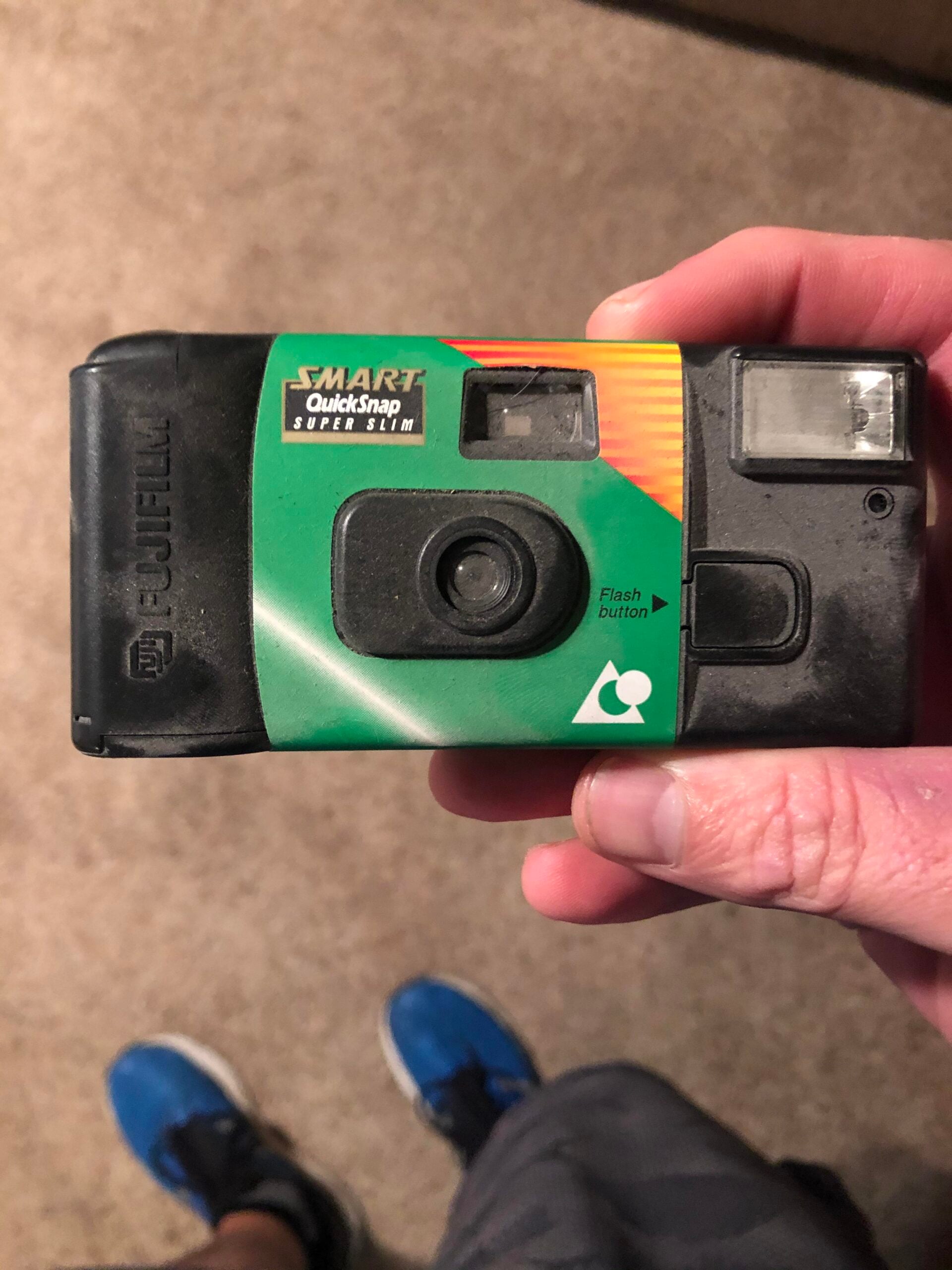 A hand holds a Fujifilm Smart QuickSnap Super Slim disposable camera, with a green label and visible flash, against a carpeted floor. Blue sneakers are partially visible below.