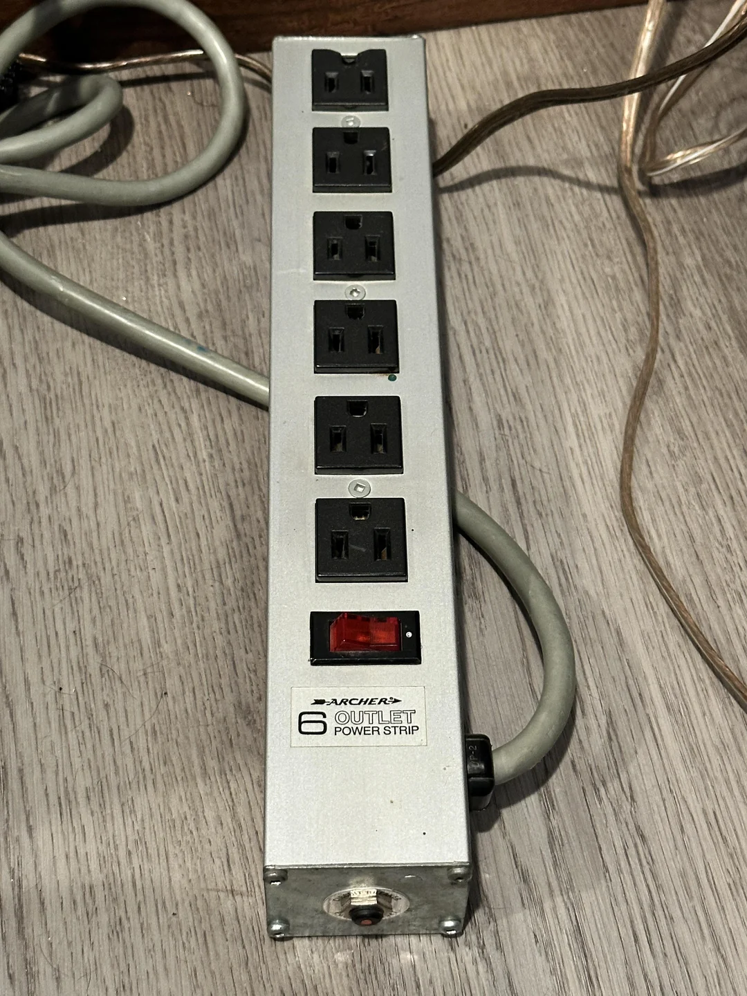 A six-outlet power strip with a red power switch and two cords plugged in, lying on a wooden floor. The power strip is labeled “Advanced 6 Outlet Power Strip.”