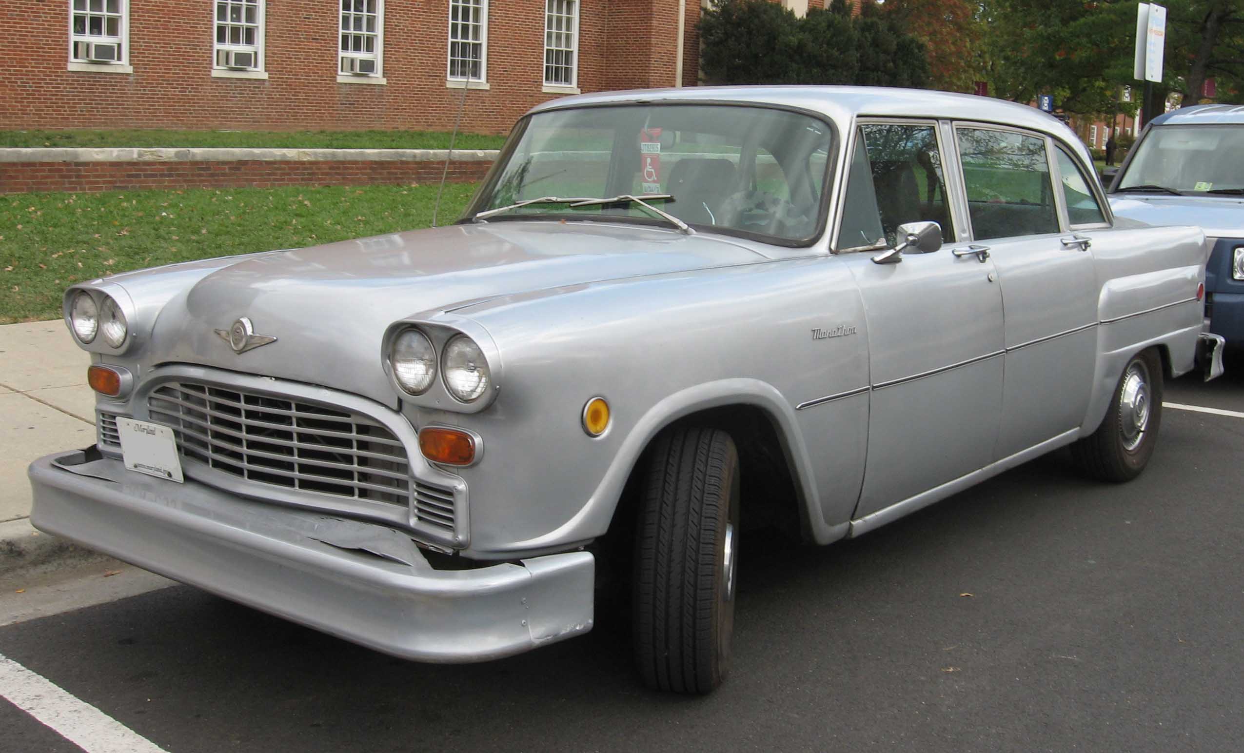 A silver vintage Checker Marathon car parked on the street near a brick building and green lawn.