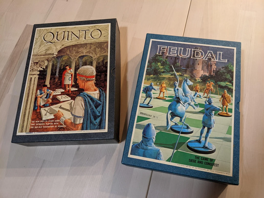 Two vintage board game boxes, “Quinto” and “Feudal,” are placed on a light wood surface. Both boxes feature illustrated medieval-themed covers with characters and game pieces.