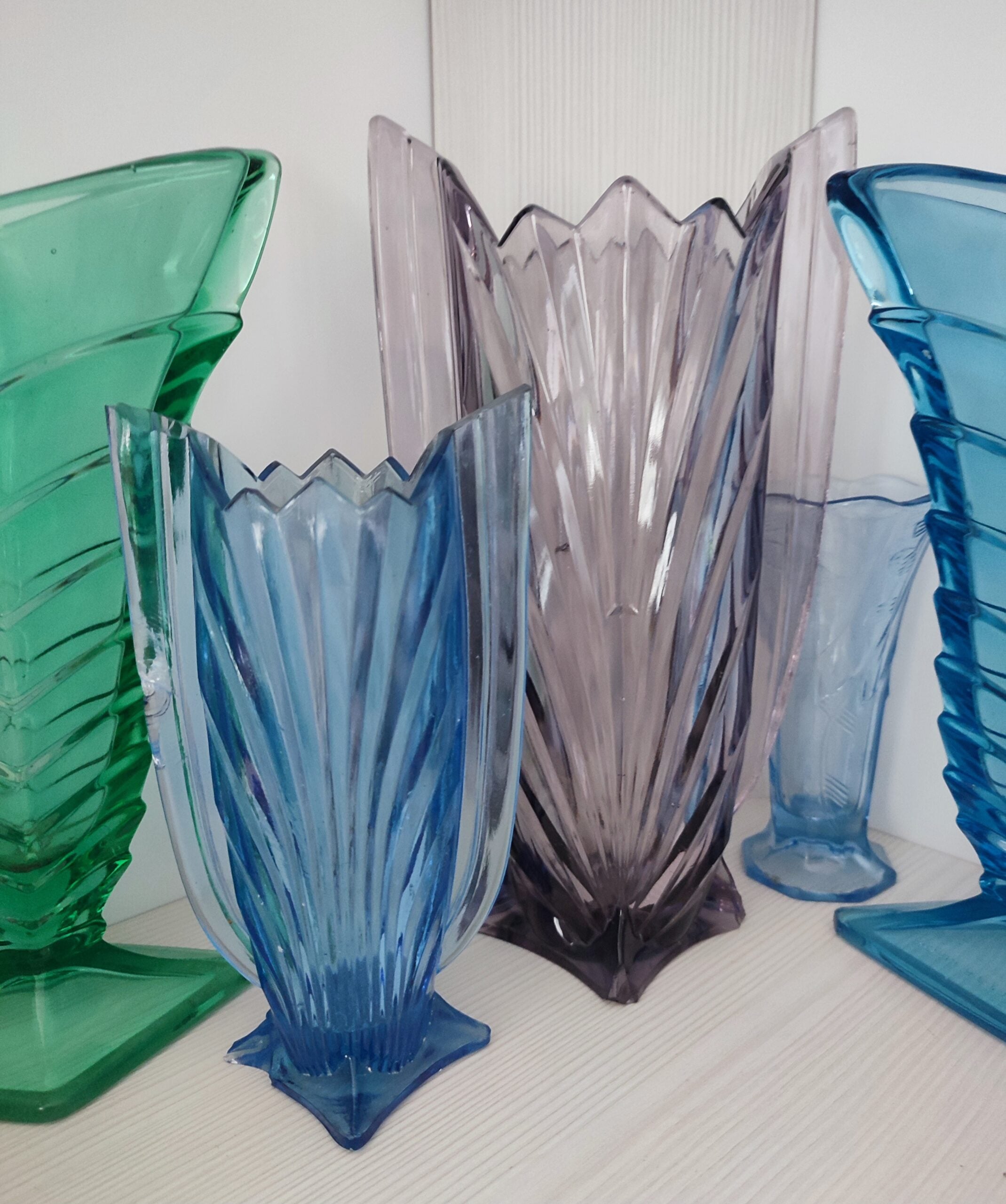 Five colorful, geometric glass vases—green, blue, purple, and turquoise—are arranged on a white wooden surface, each with unique, angular designs and vertical ridges.