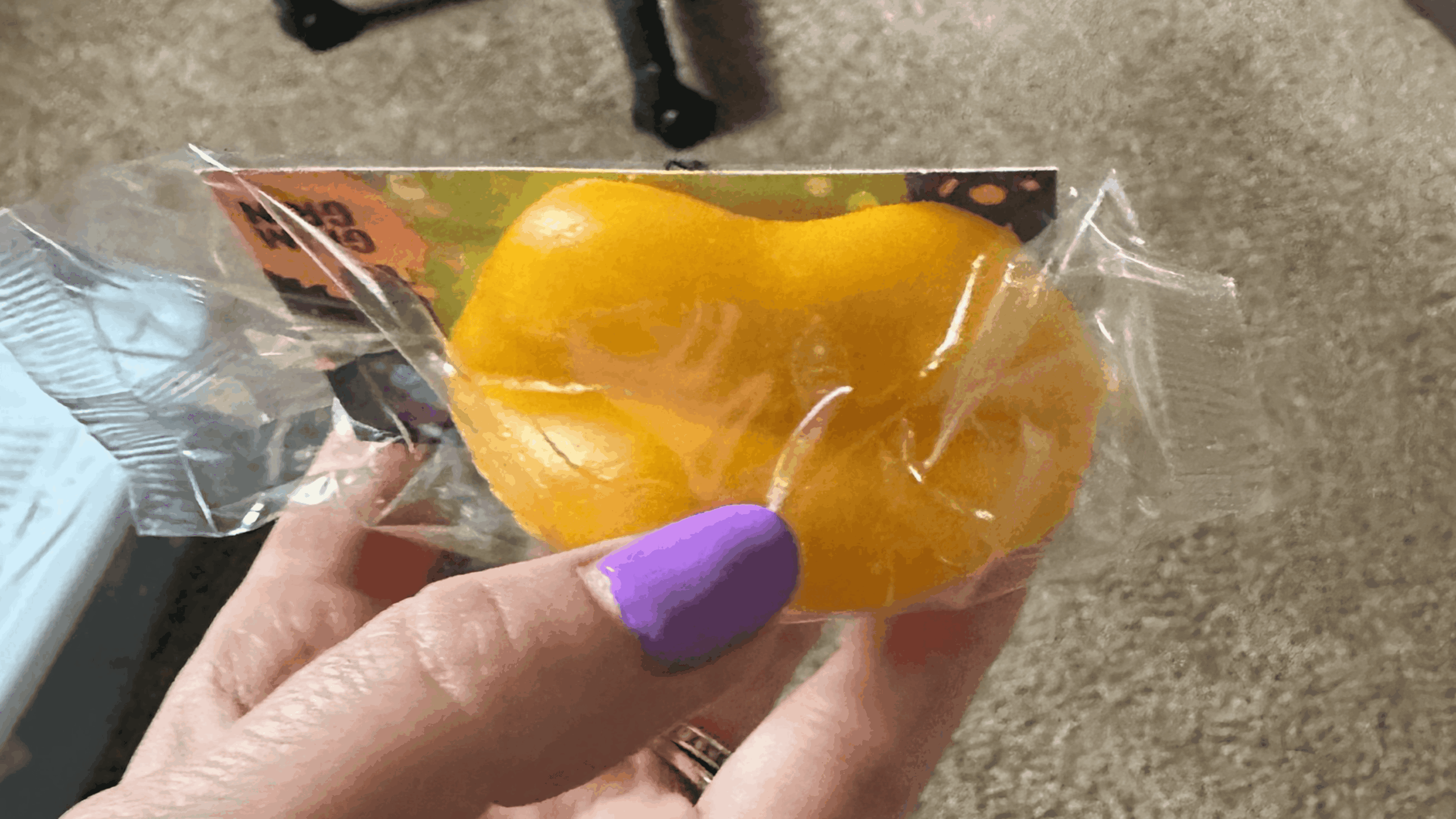 A hand with purple-painted nails holds a yellow, pumpkin-shaped object wrapped in clear plastic. The background shows a beige carpet and part of a chair.